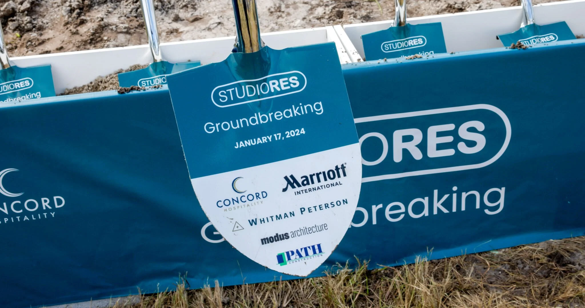 Groundbreaking ceremony with a blue banner and a sign that reads 'Groundbreaking January 17, 2024', featuring logos of Marriott, Concord Hospitality, Whitman Peterson, and other sponsors, at a dirt site.