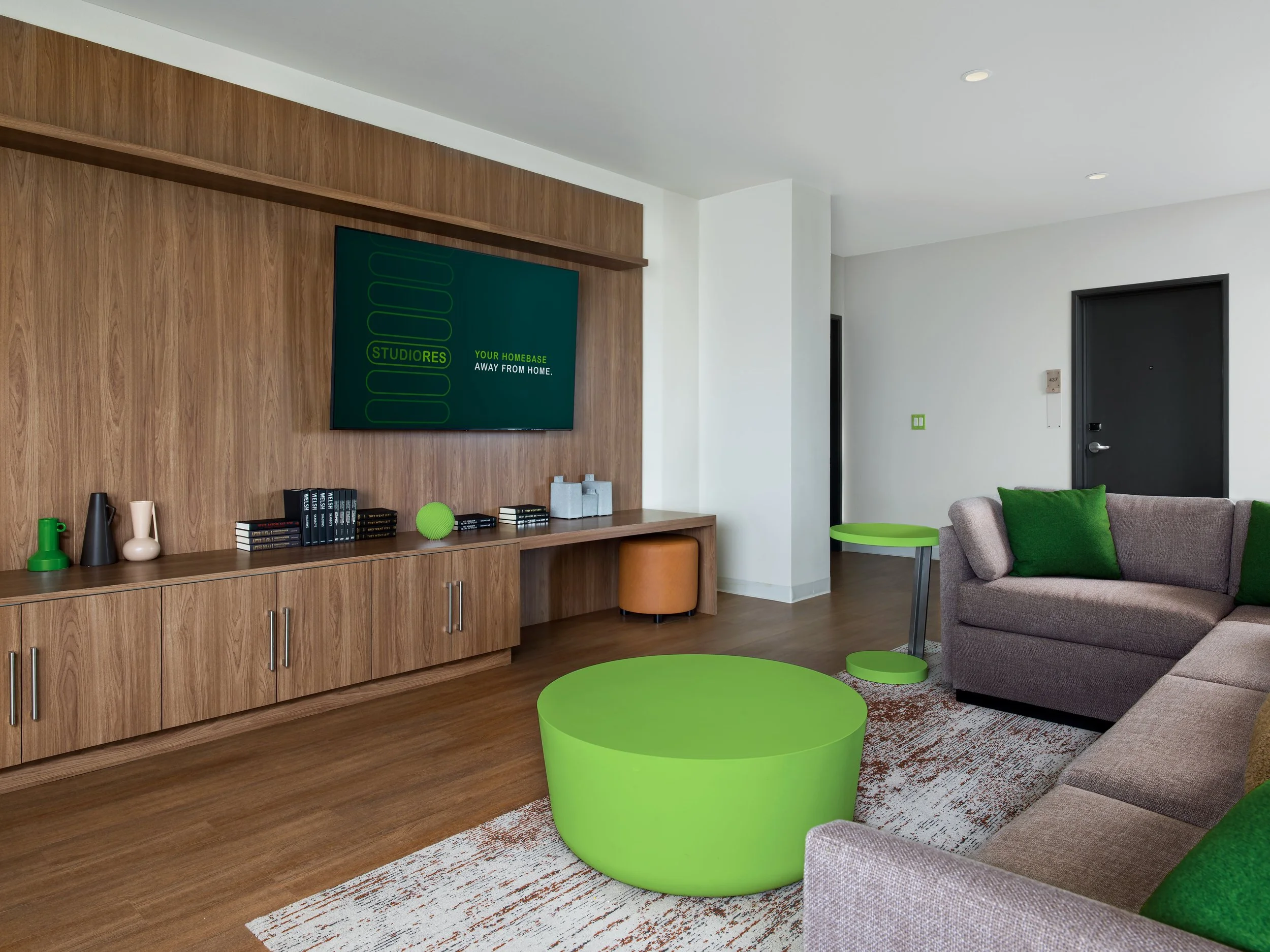 Modern living room with gray sofa and green accent pillows, lime green round coffee table, matching side table, and wooden media console, with wall-mounted TV displaying a green and black screen.