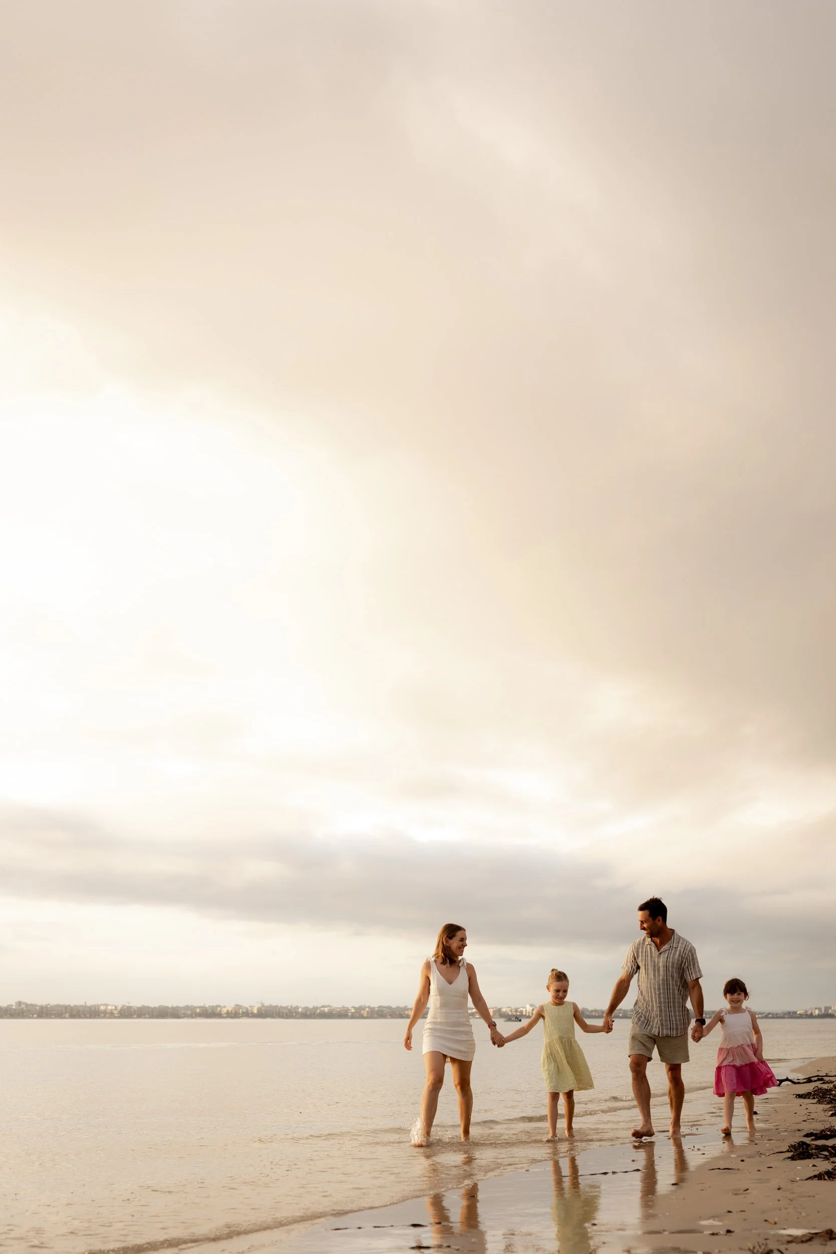 Family of four holding hands and walking through the shallow water at sunset during a Sutherland Shire beach photoshoot.