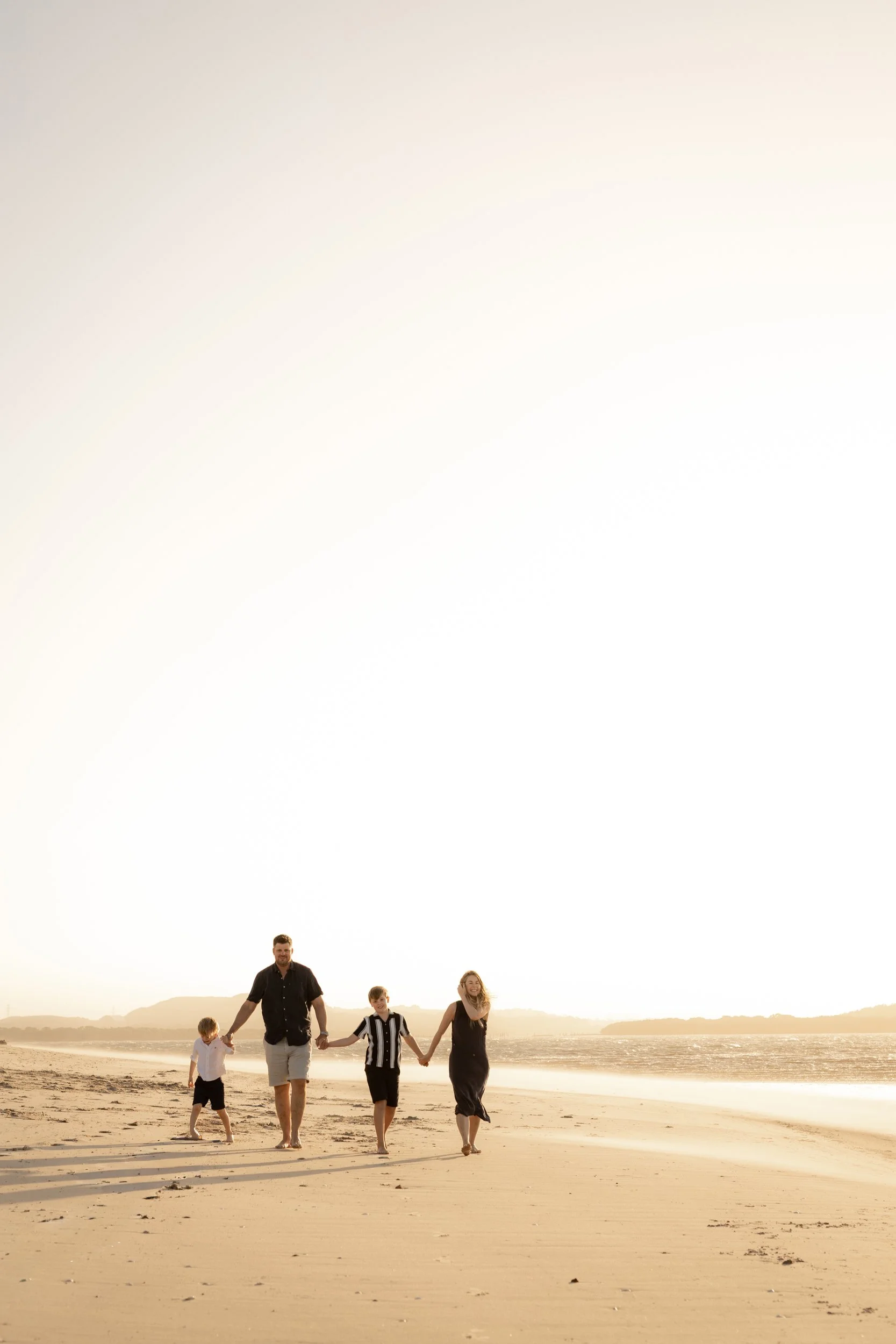 A family photo taken from afar, showing the parents and their two sons walking hand in hand over the sand during golden hour in Kurnell.