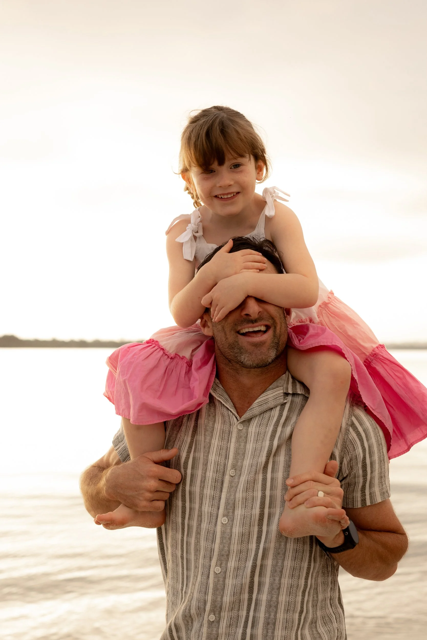 Little girl sitting on her father’s shoulders, holding his eyes shut, during a joyful family photo shoot at sunset on Kurnell beach.