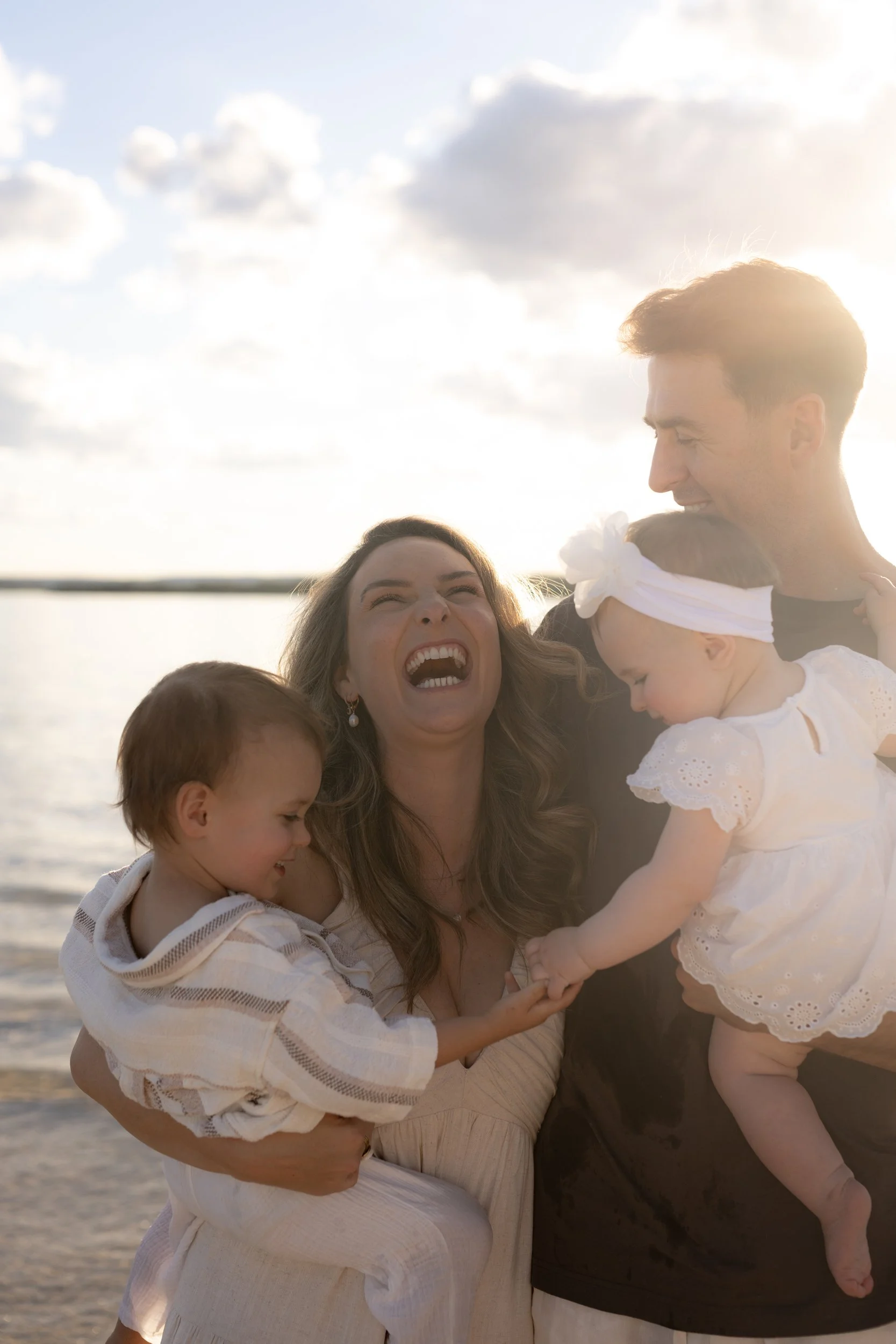 Family photo at Kurnell Beach, photographed by Captured by Jane during a Christmas Minis session. The parents are holding their two kids who are holding hands; the mum is laughing beautifully.