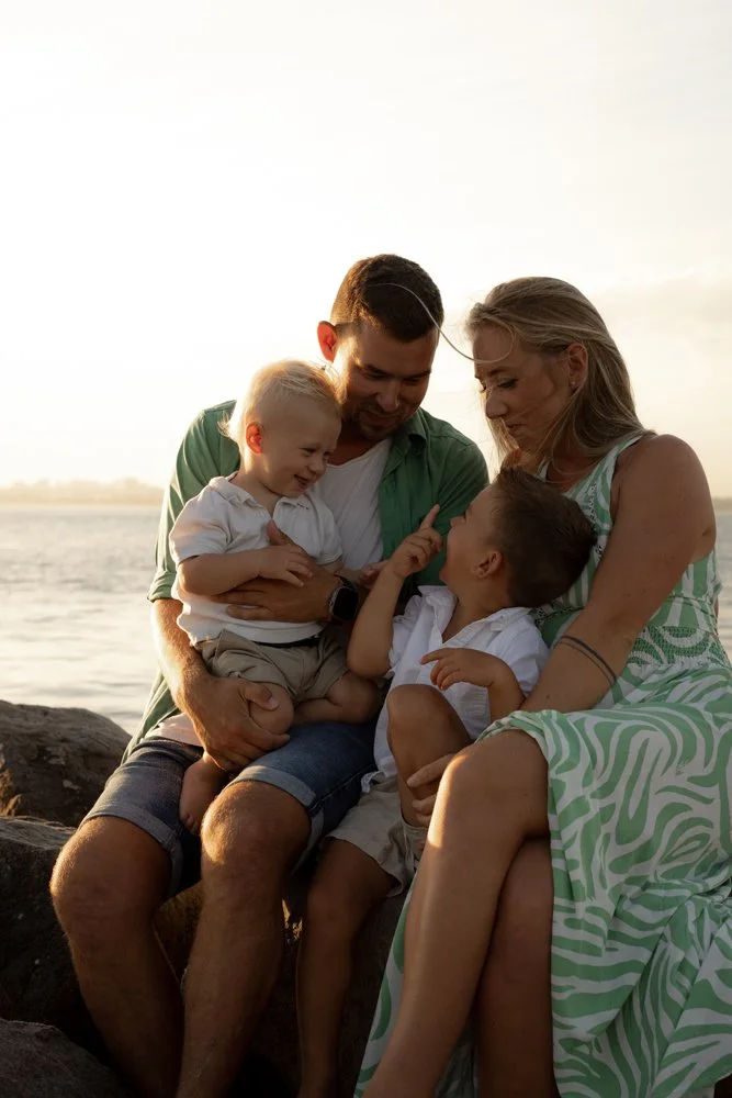 A family of four sitting on rocks near the water during sunset, smiling and engaging with each other.