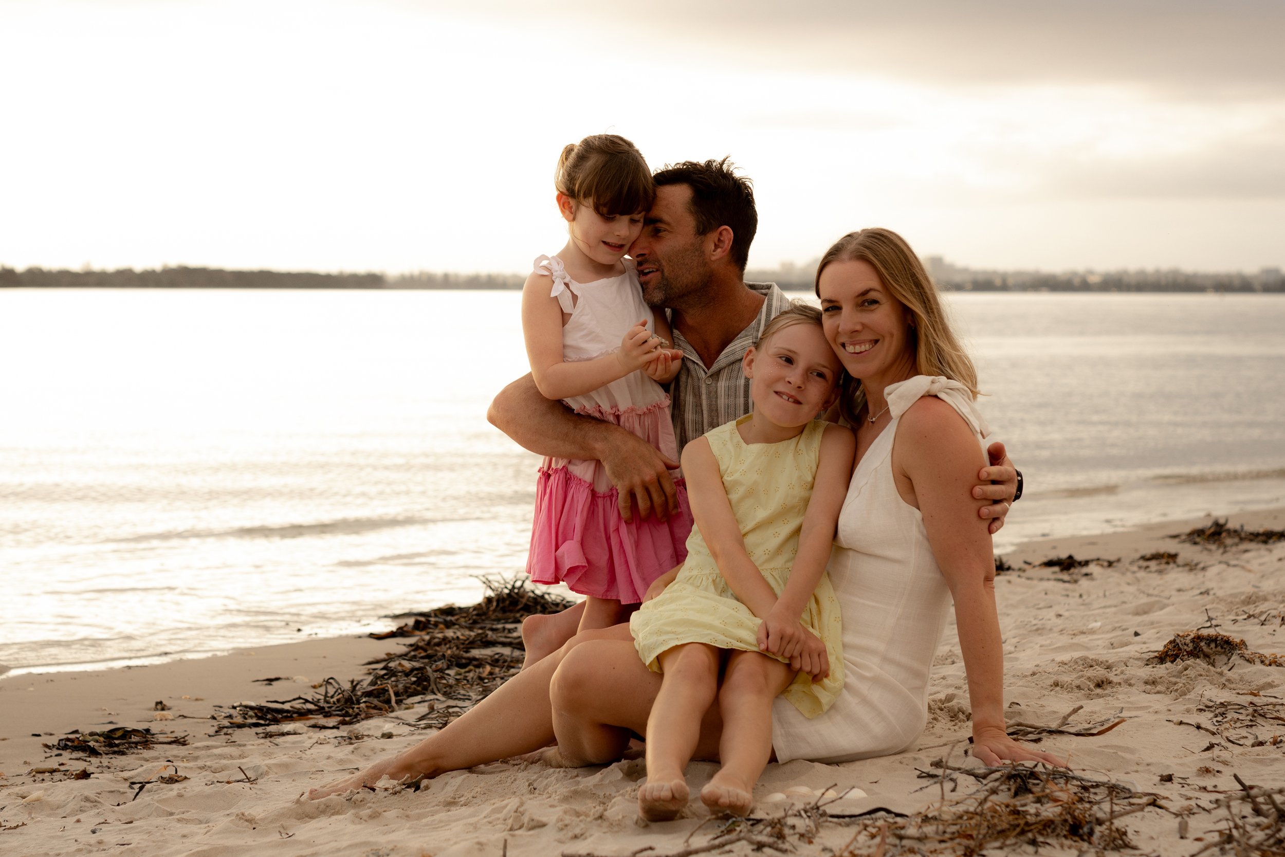 A family of four, all snuggled up, sitting on the sand during a Christmas Minis family photoshoot in Kurnell.