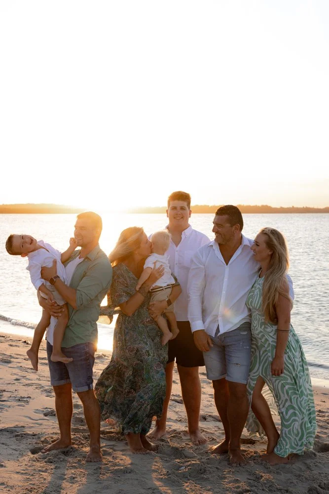 A photo of an extended family at Kurnell beach at sunset. They are wearing a mix of white, green and denim clothes, and are smiling and enjoying the moment.