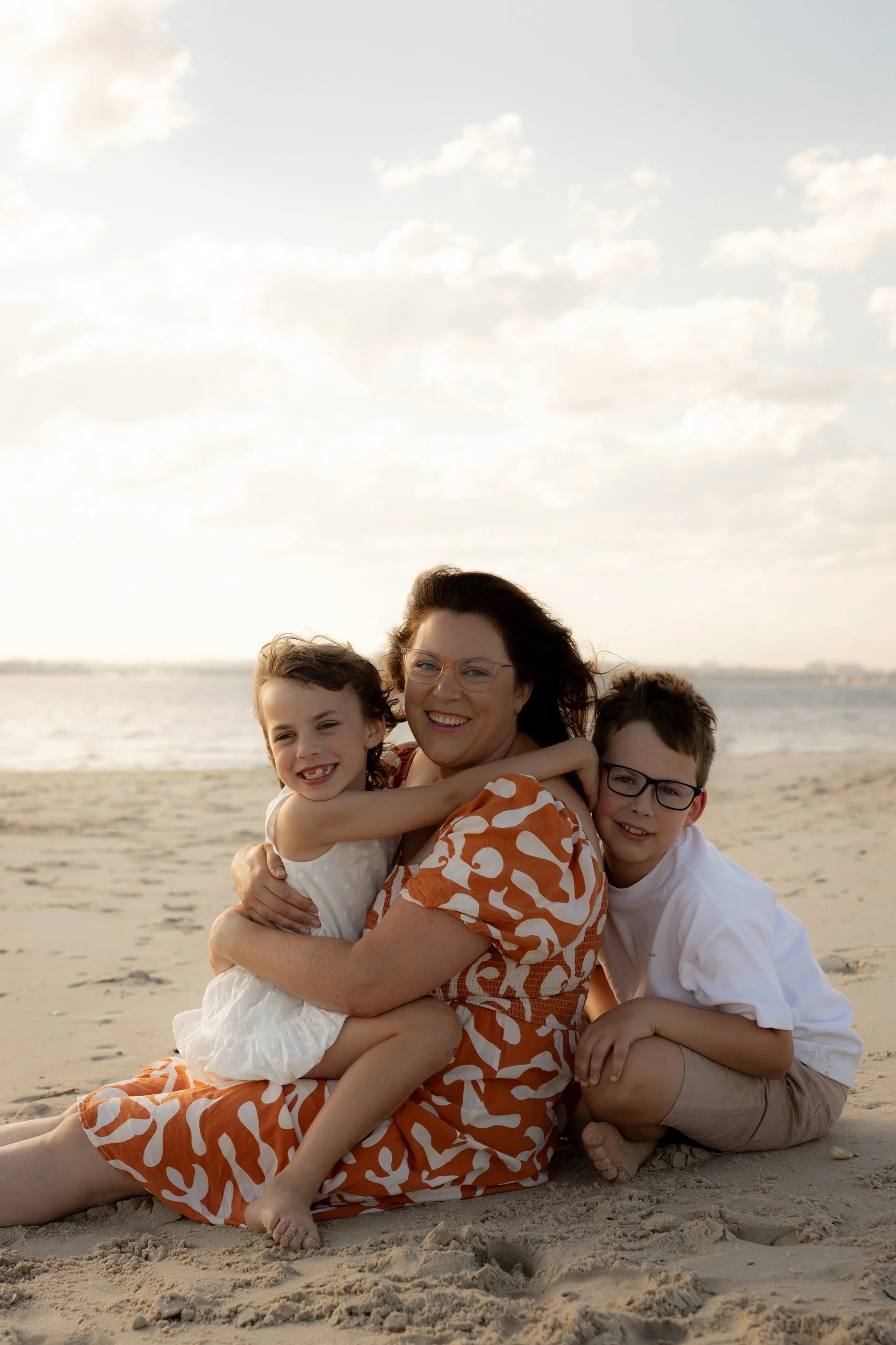 Mum sitting on the beach with her daughter in her lap and her son leaning into her from behind during a golden hour family photoshoot in Kurnell. 
