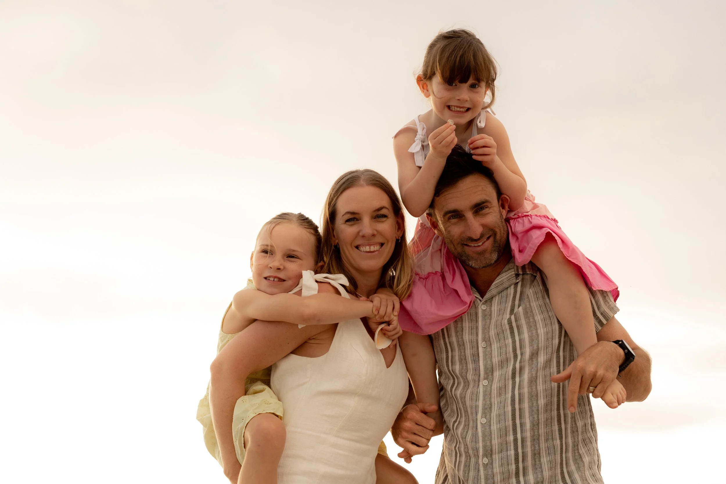 Happy family of four with the two young daughters piggy back on their parents during a Christmas beach photoshoot in the Sutherland Shire.
