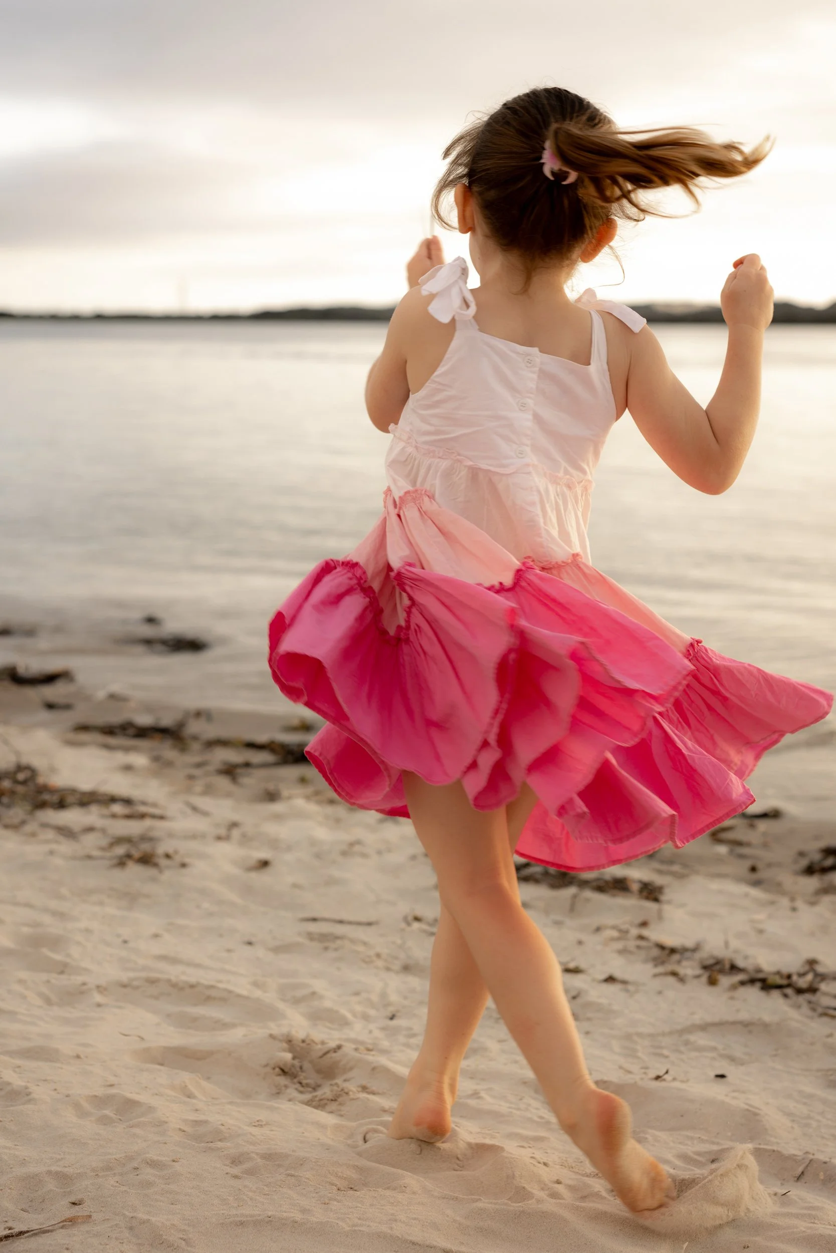 Little girl in a flowing pink dress twirling on the beach during a playful Christmas mini photography session.