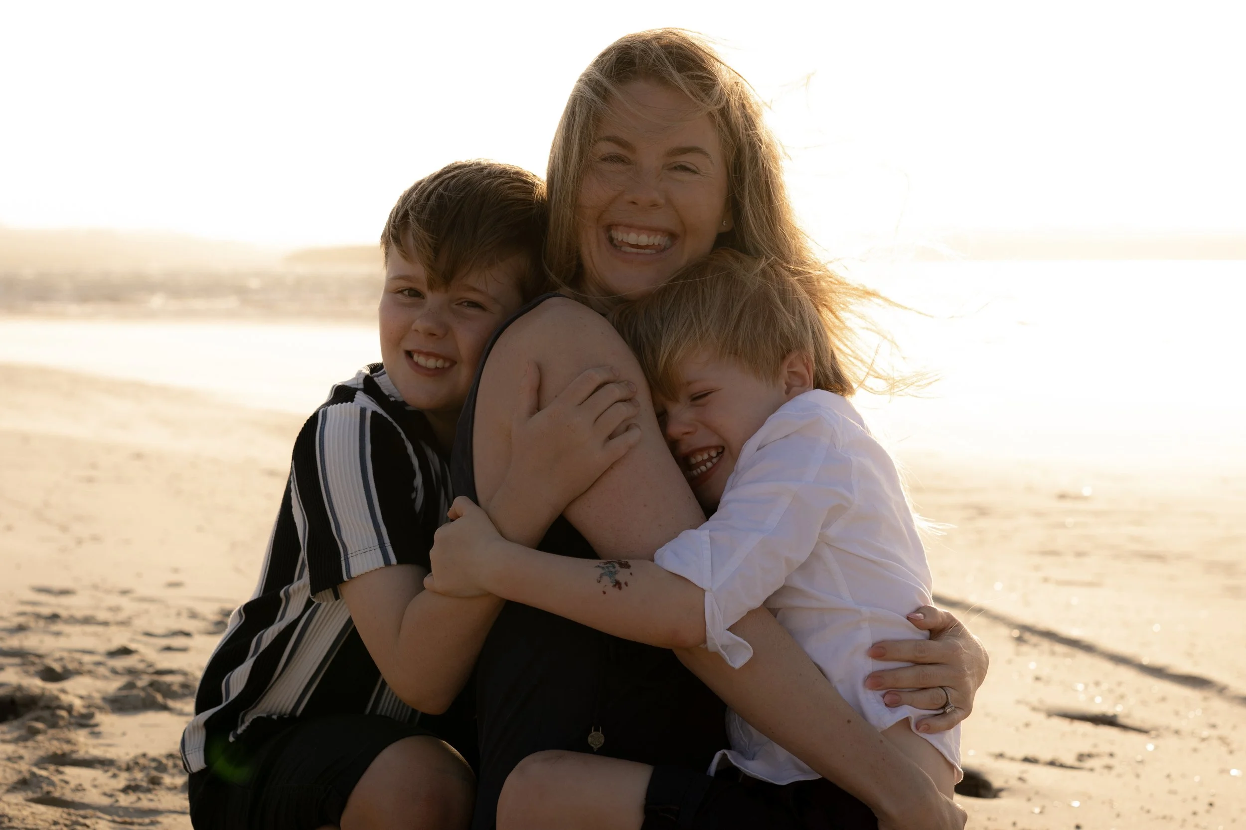 Mother laughing as her two sons hug her on the beach during a fun Christmas mini family session near Cronulla.