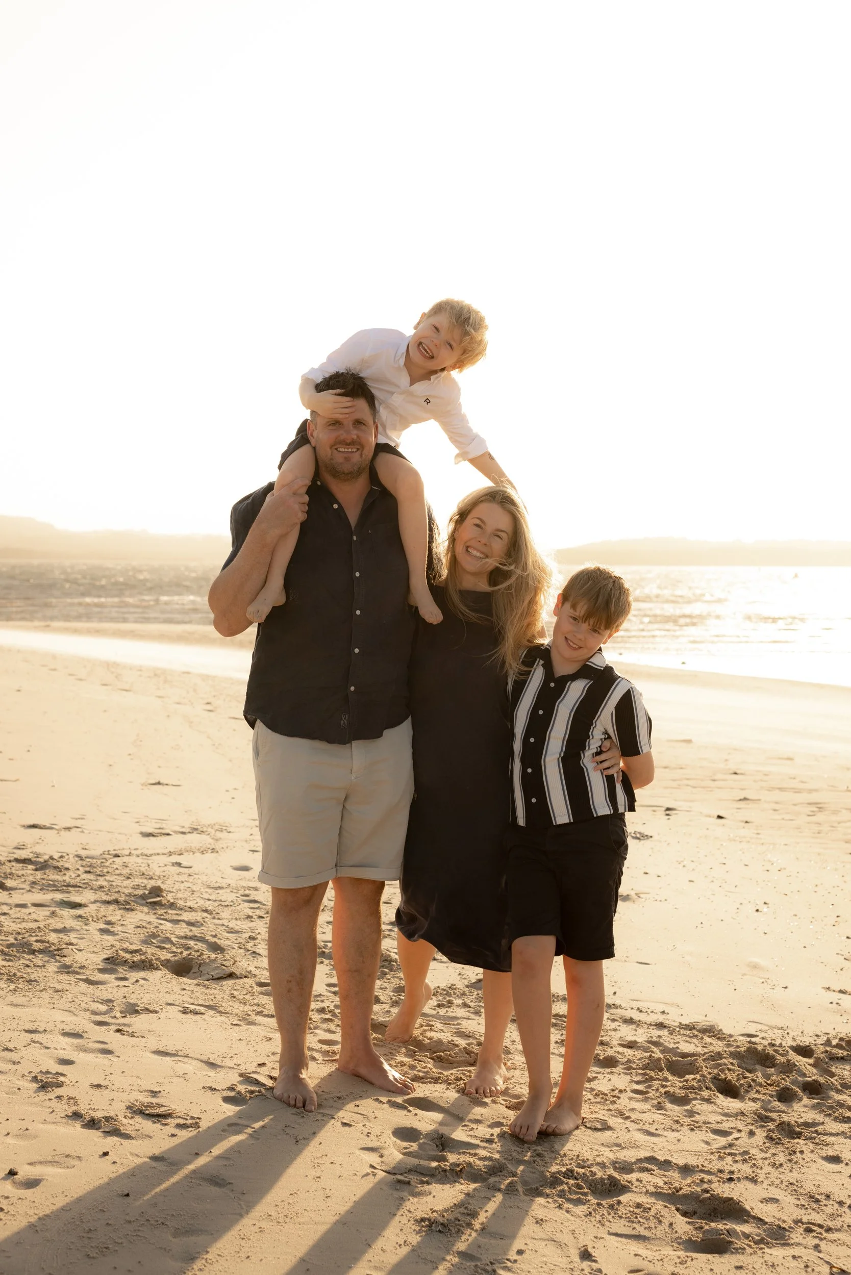 Family of four laughing together during a sunset beach photoshoot in the Sutherland Shire. The youngest sits on dads shoulders and teases mums hair from above.