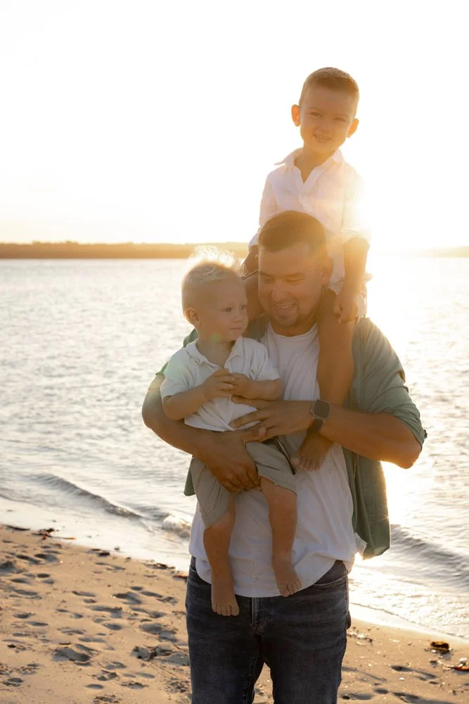 A father holding his two young boys at Kurnell beach at sunset. One of the boys is sitting on his shoulders, the other is in his arms, they are all smiling.