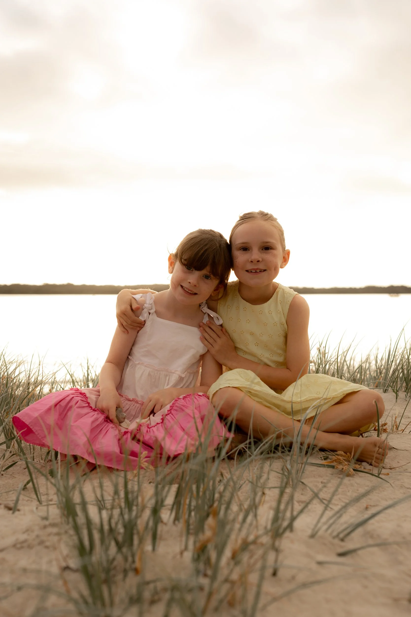 Two sisters in colourful dresses sitting in the sanddunes of Kurnell during golden hour, smiling at the camera. 