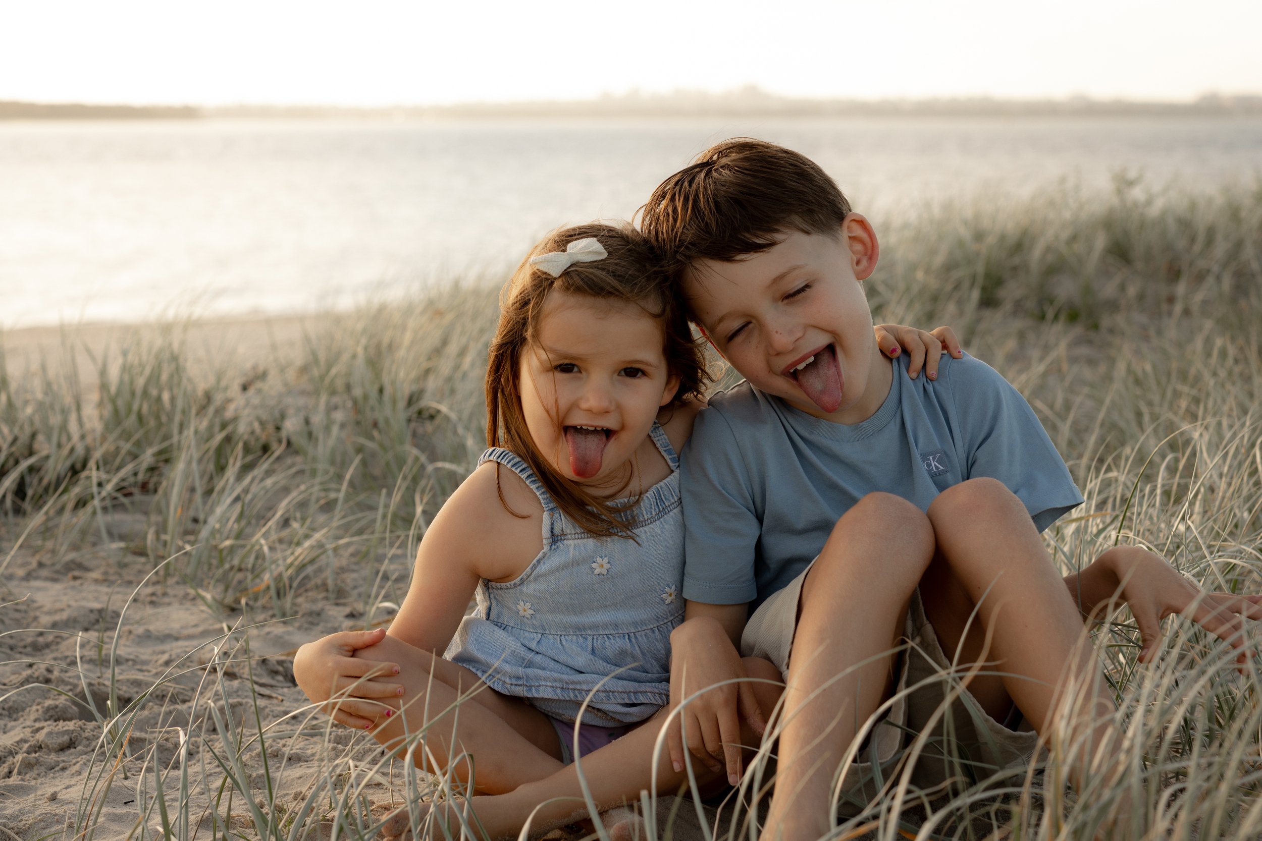 Two cheeky kids, sitting in the beach grass with the ocean in the background, both pointing their tongues at the photographer.