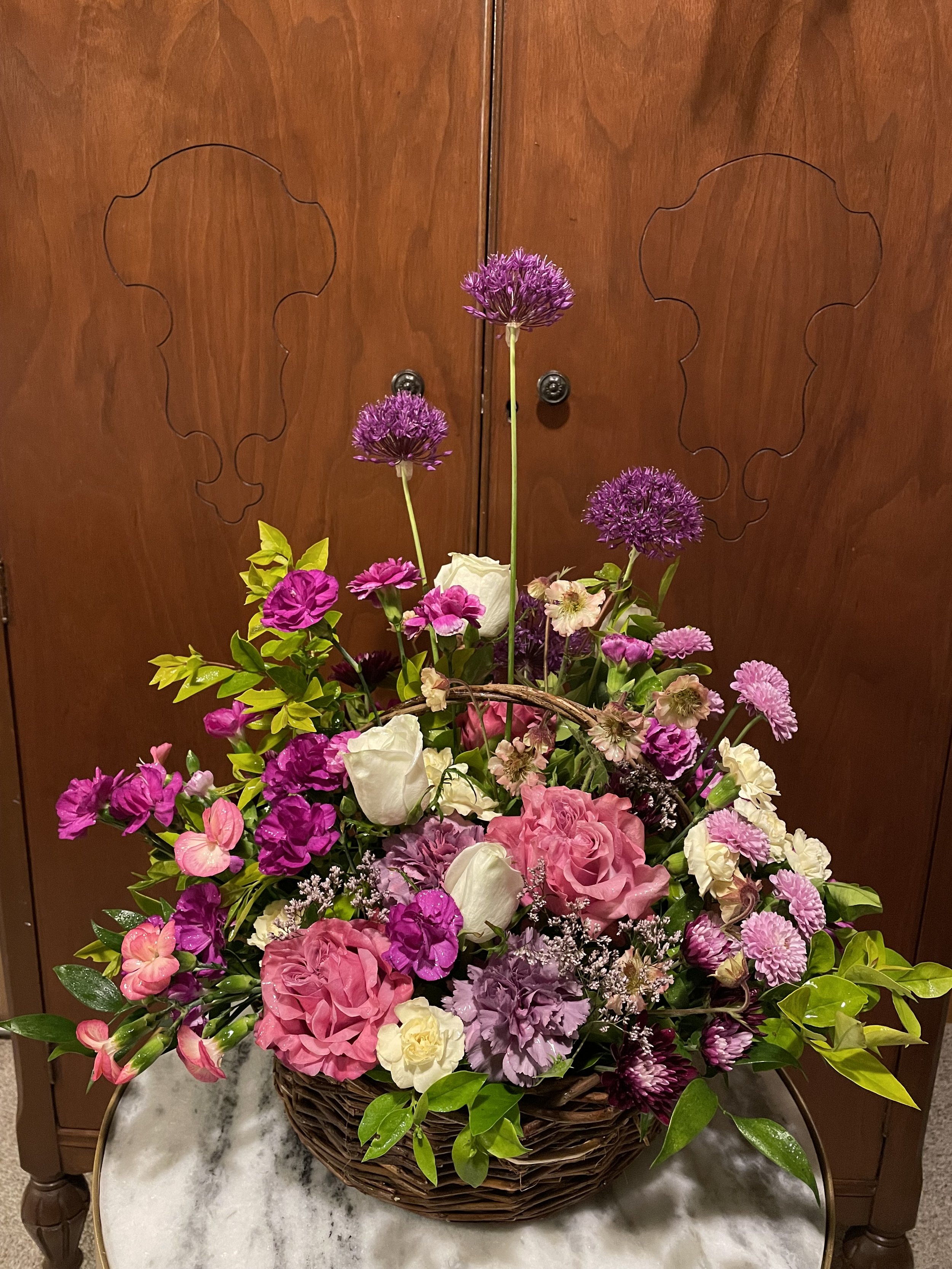 A basket of assorted pink, purple, and white flowers on a marble table in front of a wooden cabinet.