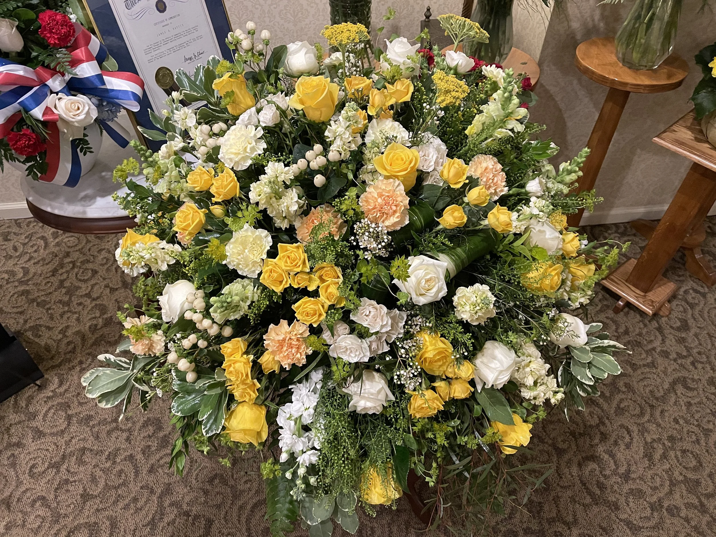 Large floral arrangement with white, yellow, and light pink flowers, including roses and carnations, on a brown table with wood stools on either side, in a room with framed certificates or awards on the wall.