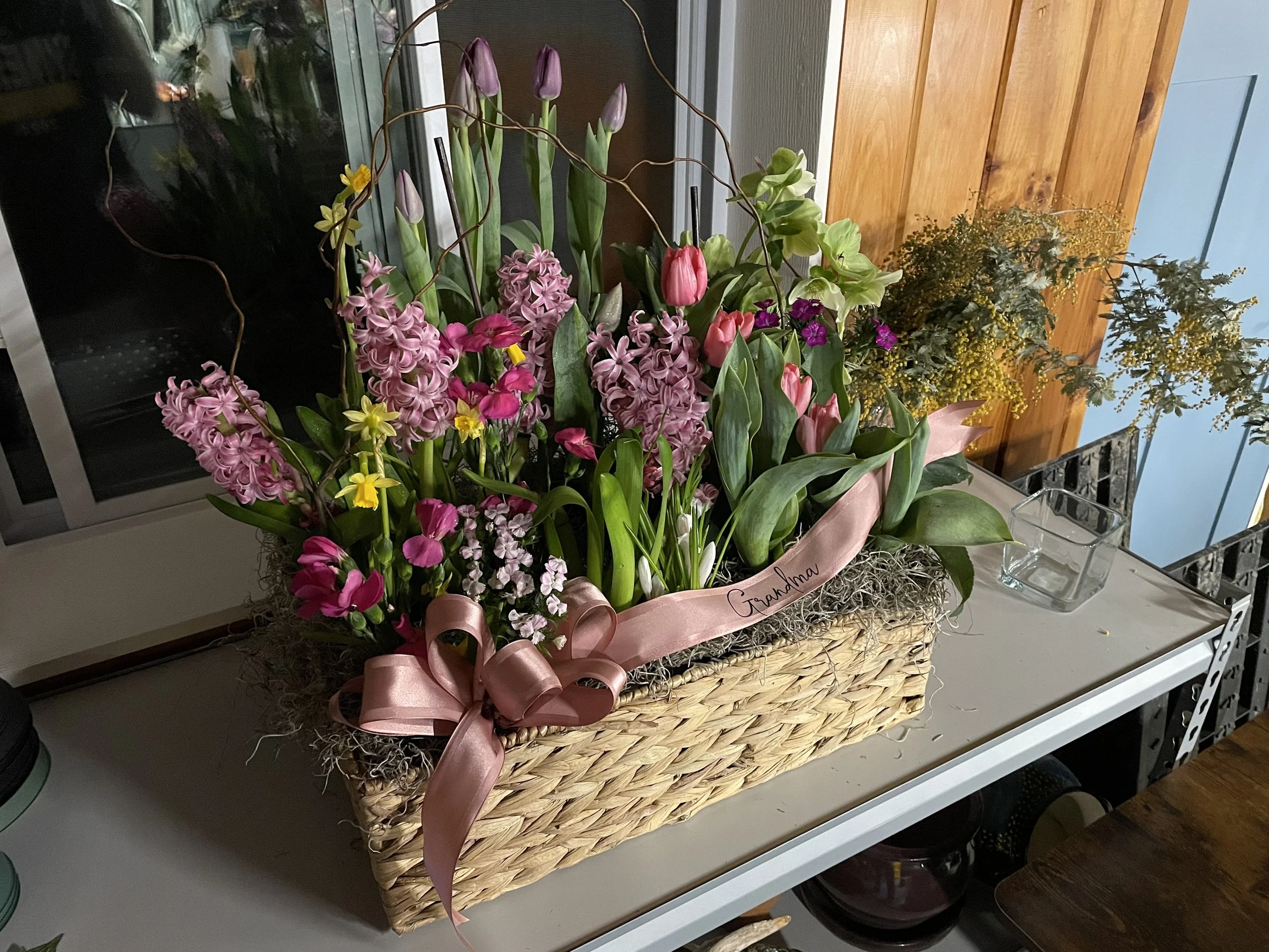 Arrangement of pink, purple, and yellow flowers in a woven basket with a pink ribbon and green foliage, placed on a white table.