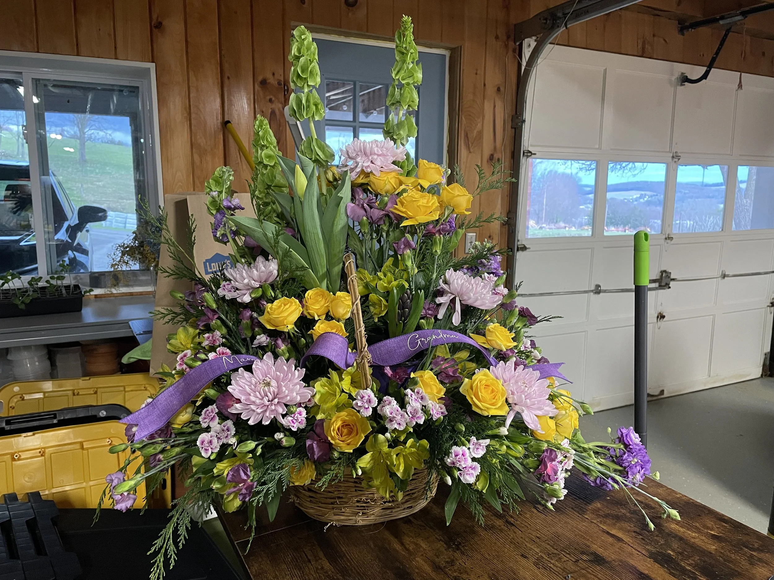 A colorful flower arrangement in a basket on a wooden table in a garage, with a purple ribbon that says 'Grandma' and 'Mom' tied around it.