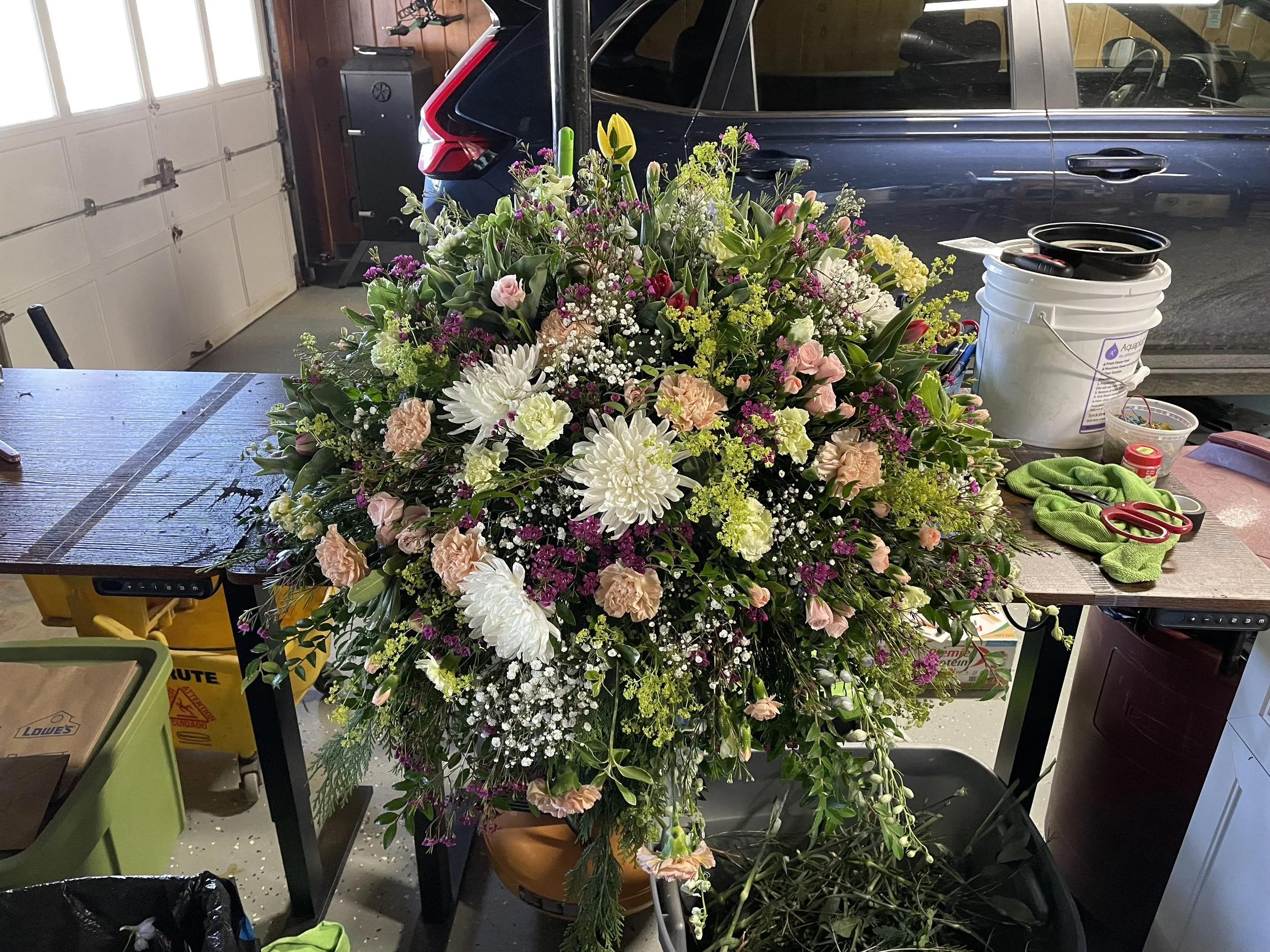 A large, colorful flower arrangement in a brown pot in a garage, with a black car and various gardening and floral tools on a wooden table.