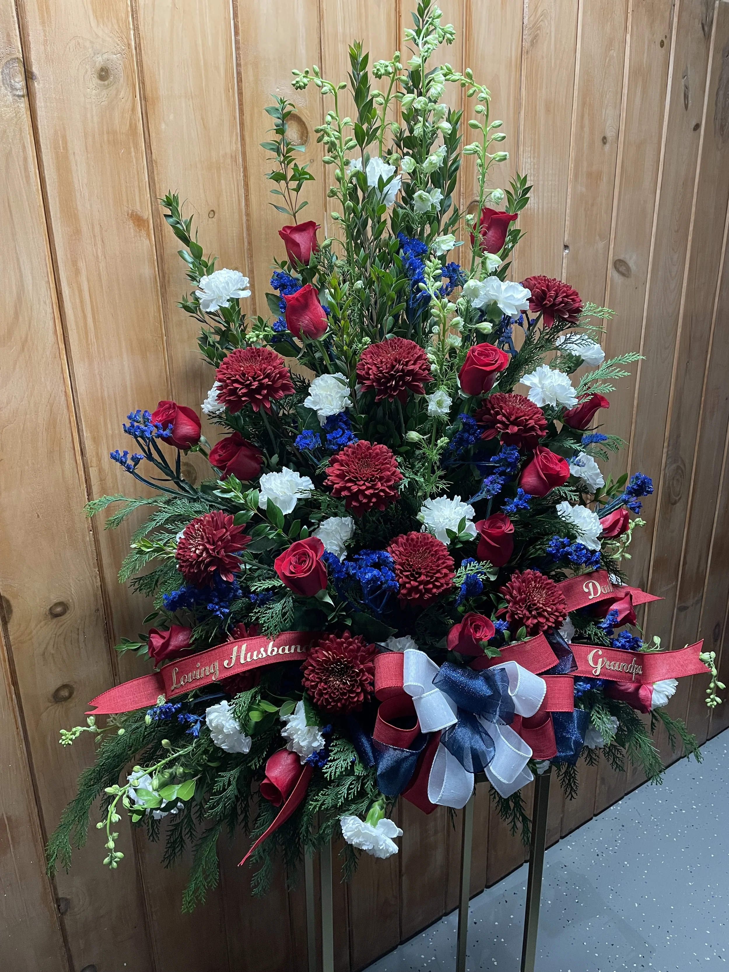 A large floral arrangement with red, white, and blue flowers, including roses and chrysanthemums, on a stand against a wooden wall. It has a ribbon with a message dedicated to a loved one.