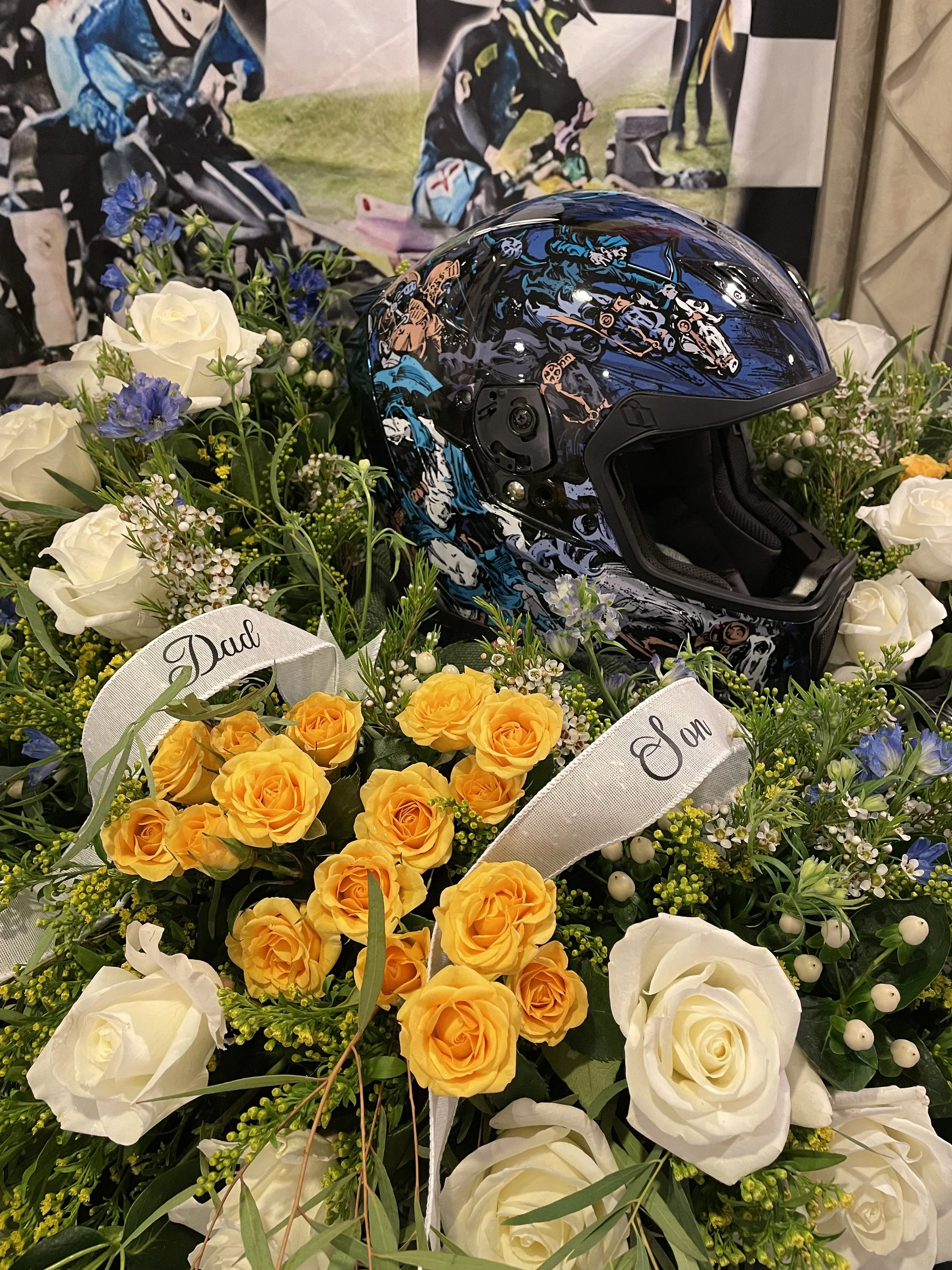 A black motorcycle helmet with colorful racing graphics sits on a flower arrangement that includes white and yellow roses, small white and blue flowers, and greenery. There are white ribbons labeled "Dad" and "Son" among the flowers.