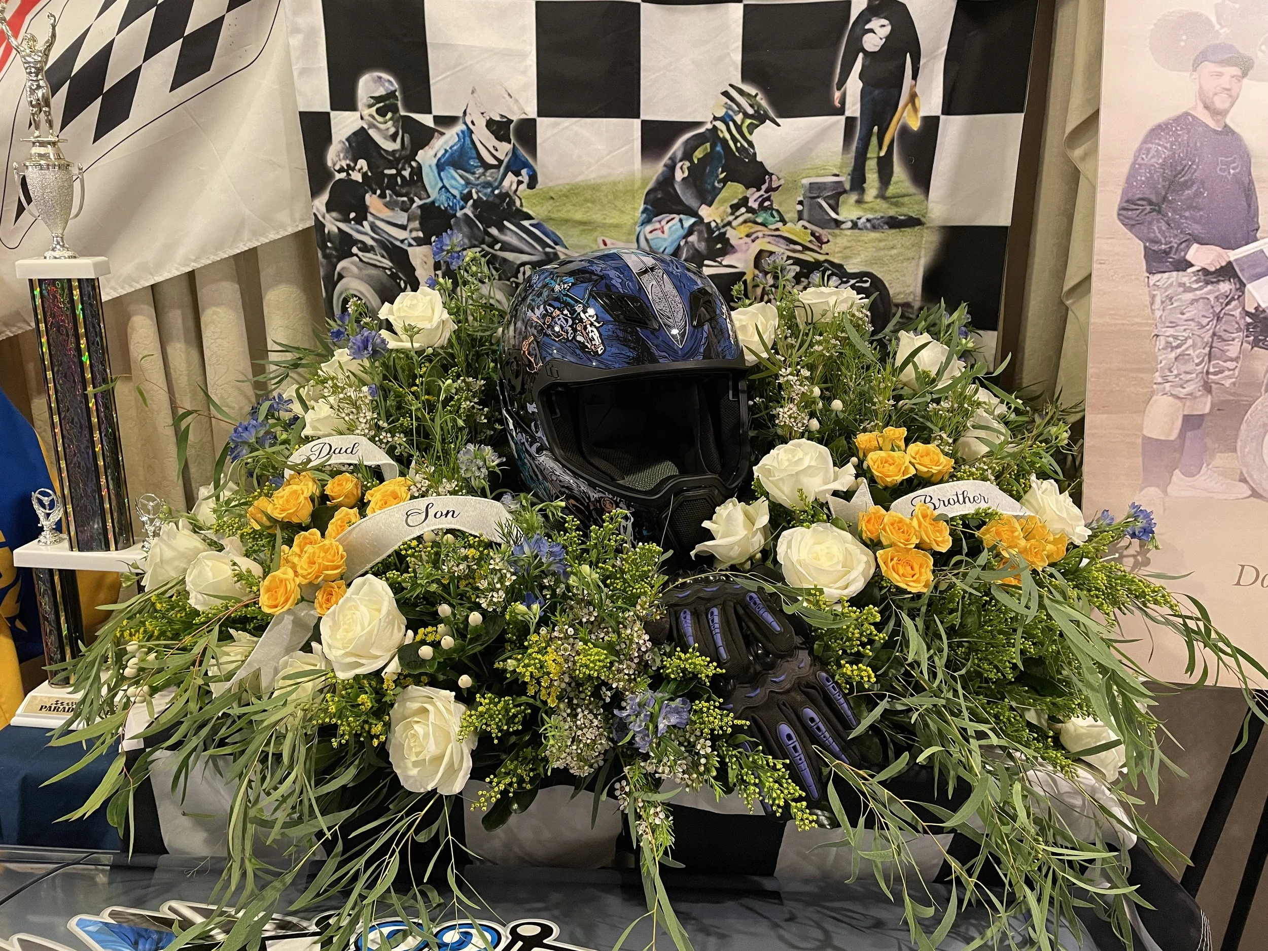 A memorial display with a motorcycle helmet, black gloves, and a floral arrangement of white and yellow roses, white lilies, and greenery. Ribbons with labels read 'Dad' and 'Son,' and a motocross picture as a backdrop.
