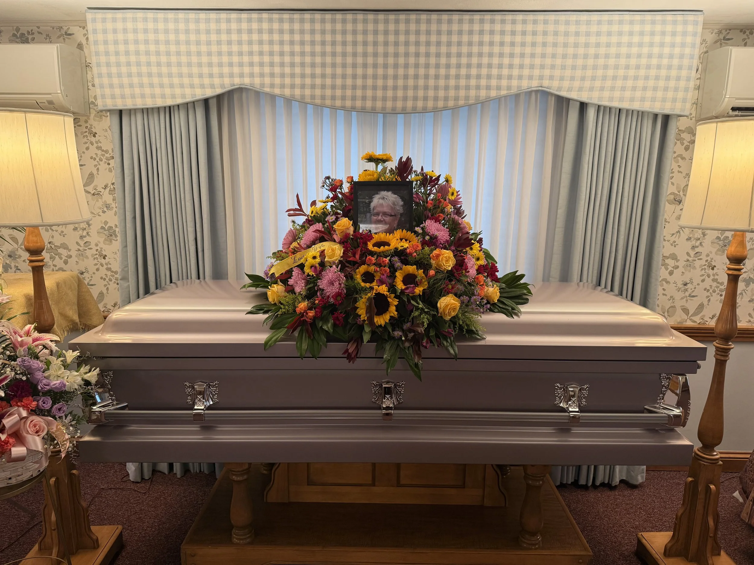 Coffin with floral arrangements and a framed photo of an elderly woman, set in a funeral parlor with floral wallpaper, curtains, and lamps.