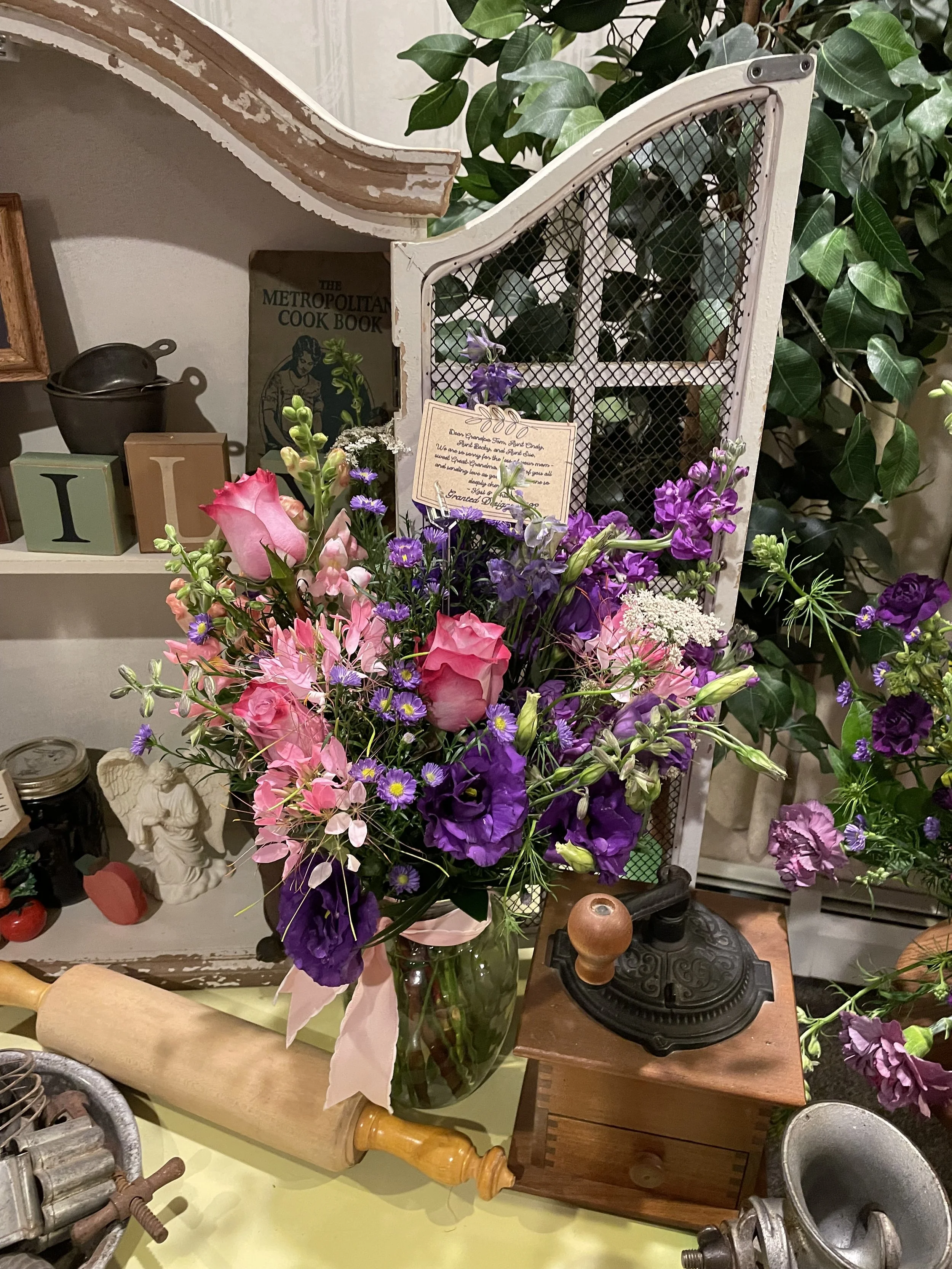A bouquet of pink, purple, and white flowers in a glass vase on a yellow surface, with a rolling pin and vintage kitchen tools nearby. In the background, there are decorative items and plants.
