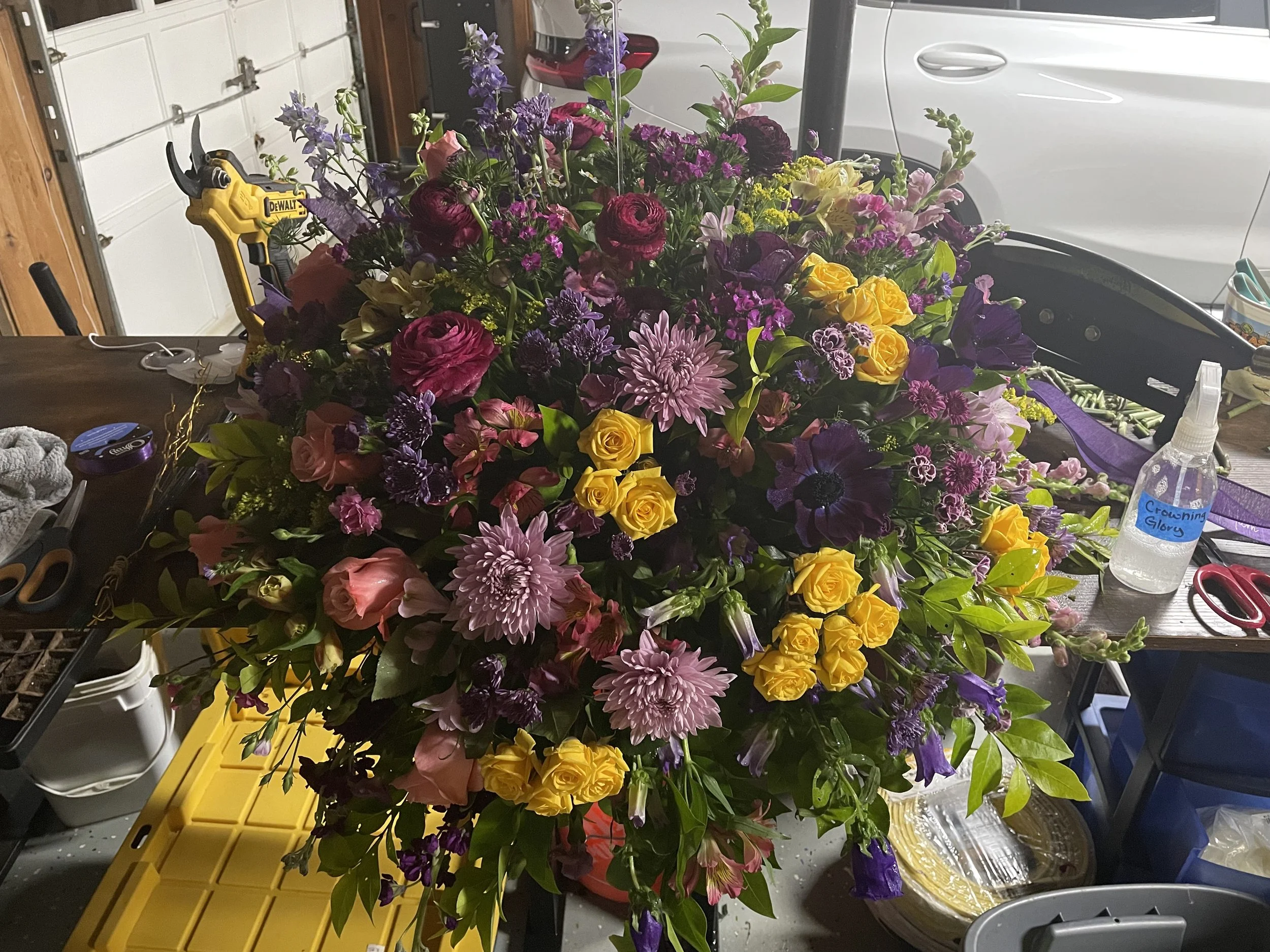 Large colorful flower arrangement on a work table, including purple, pink, yellow, and white flowers with green leaves, surrounded by tools and supplies in a garage or workshop.