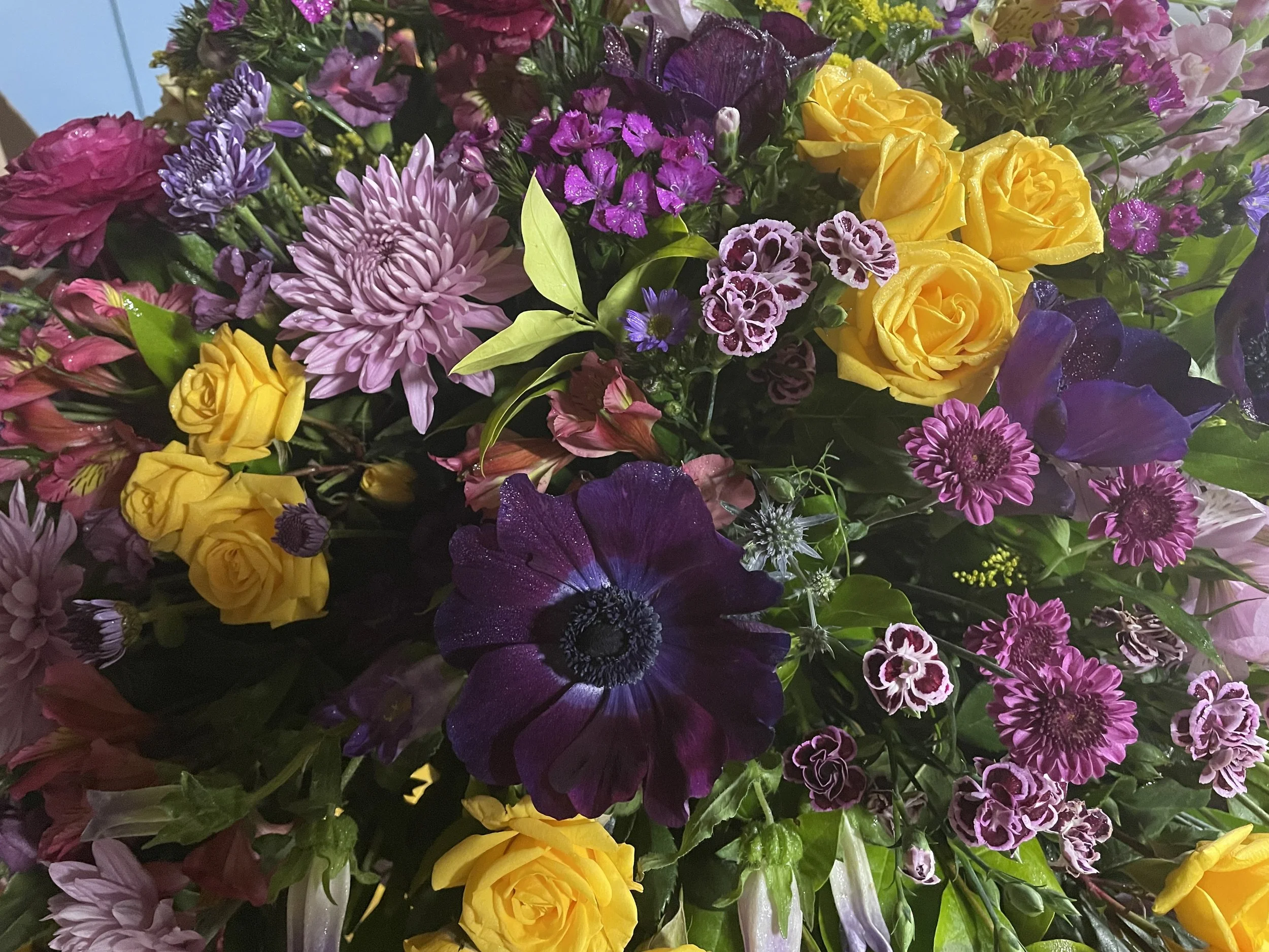A colorful bouquet of various flowers including yellow roses, purple anemones, pink chrysanthemums, and other small purple, pink, and white flowers.