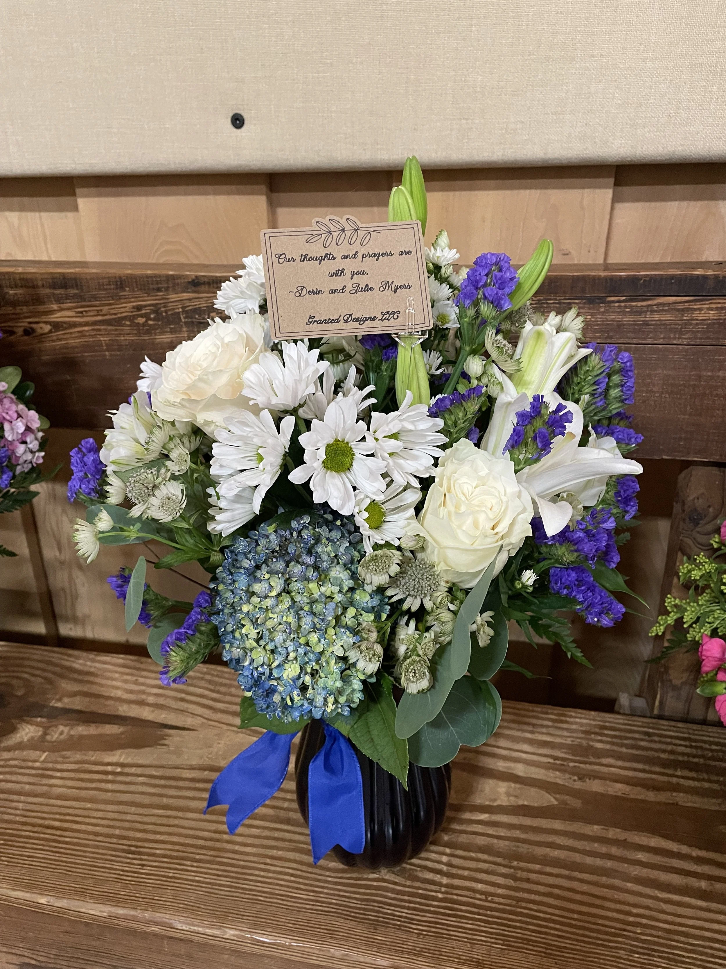 A bouquet of white, purple, and blue flowers with a blue ribbon in a black vase on a wooden table.
