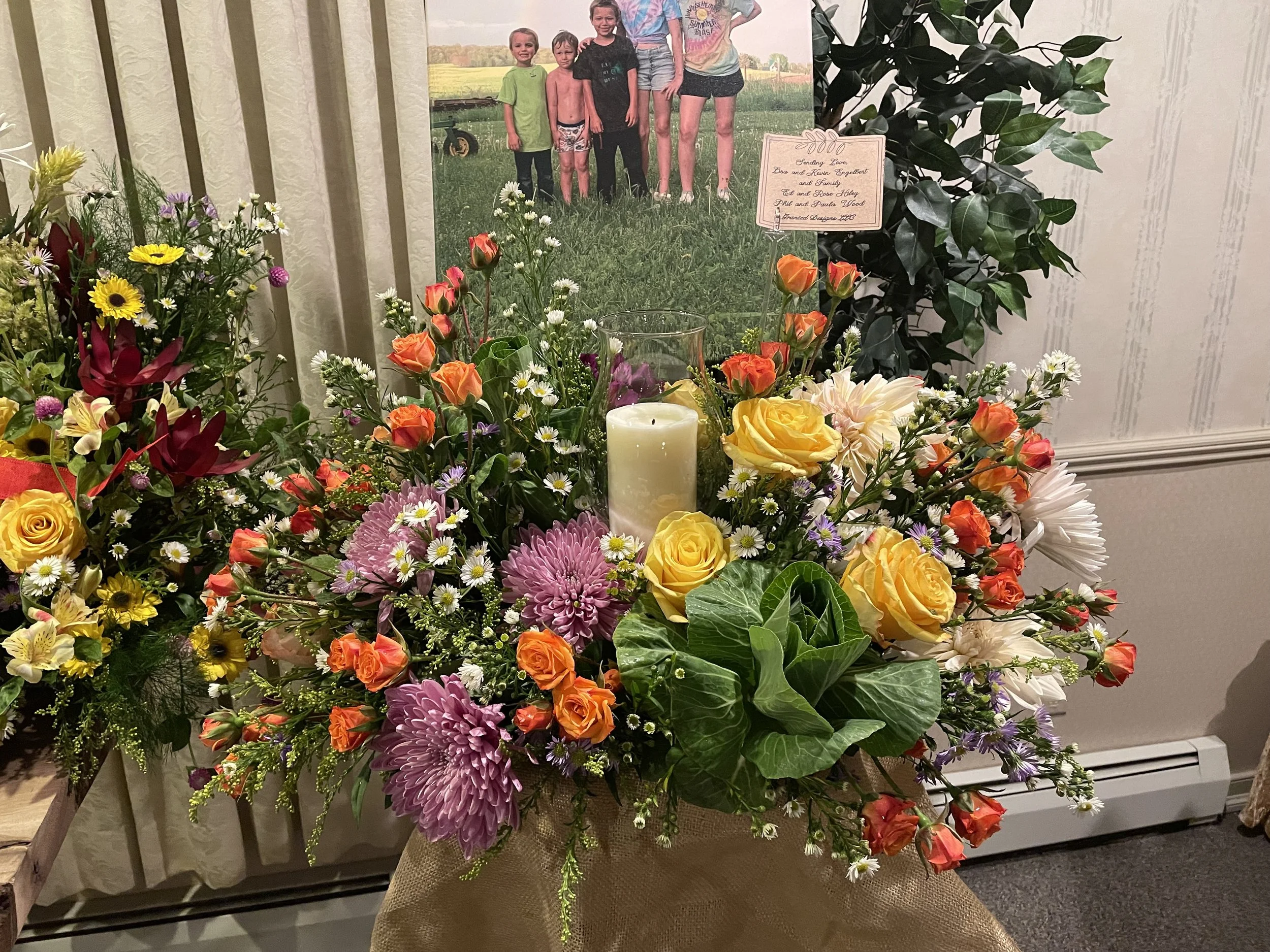 A colorful arrangement of flowers including yellow roses, purple chrysanthemums, orange spray roses, white daisies, and greenery, with a white candle in the center. In the background, there is a framed photograph of children outdoors and a card with