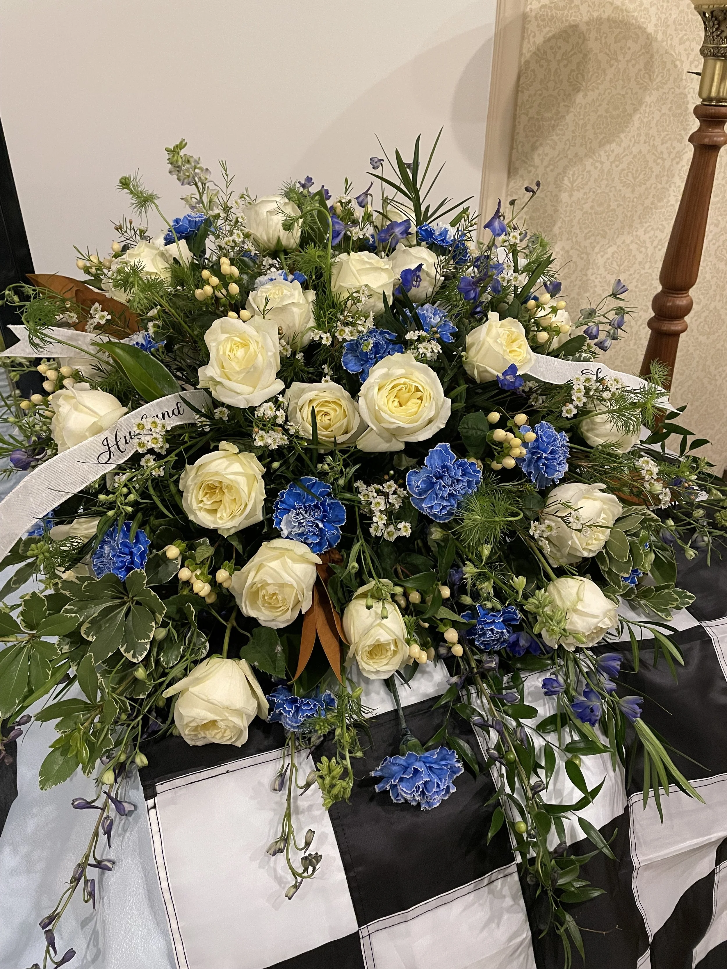 A floral arrangement with white roses, blue carnations, small white daisies, and greenery on a black and white checkered tablecloth.