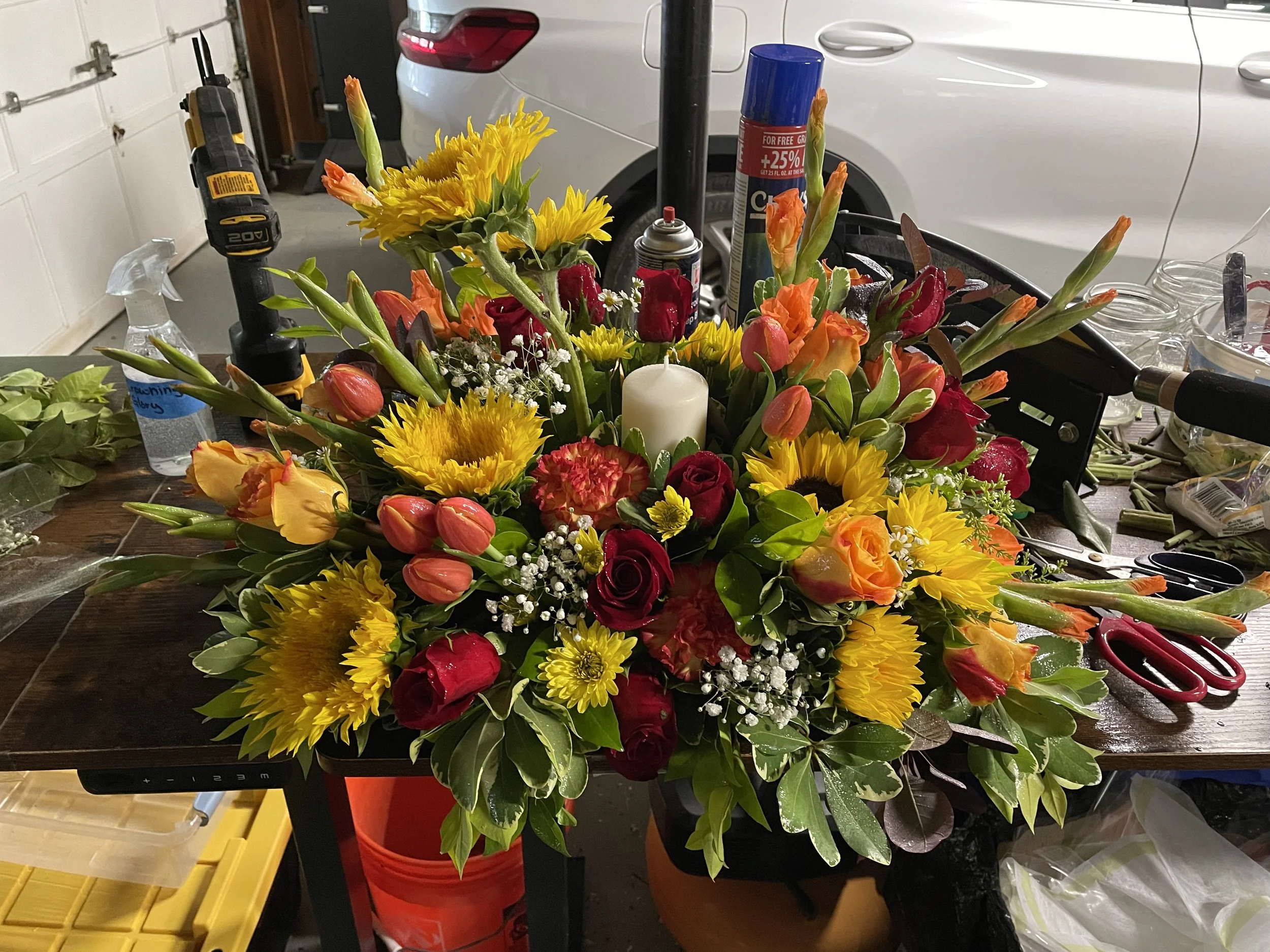 A colorful flower bouquet with sunflowers, roses, tulips, and other flowers on a workshop table, among tools and supplies.