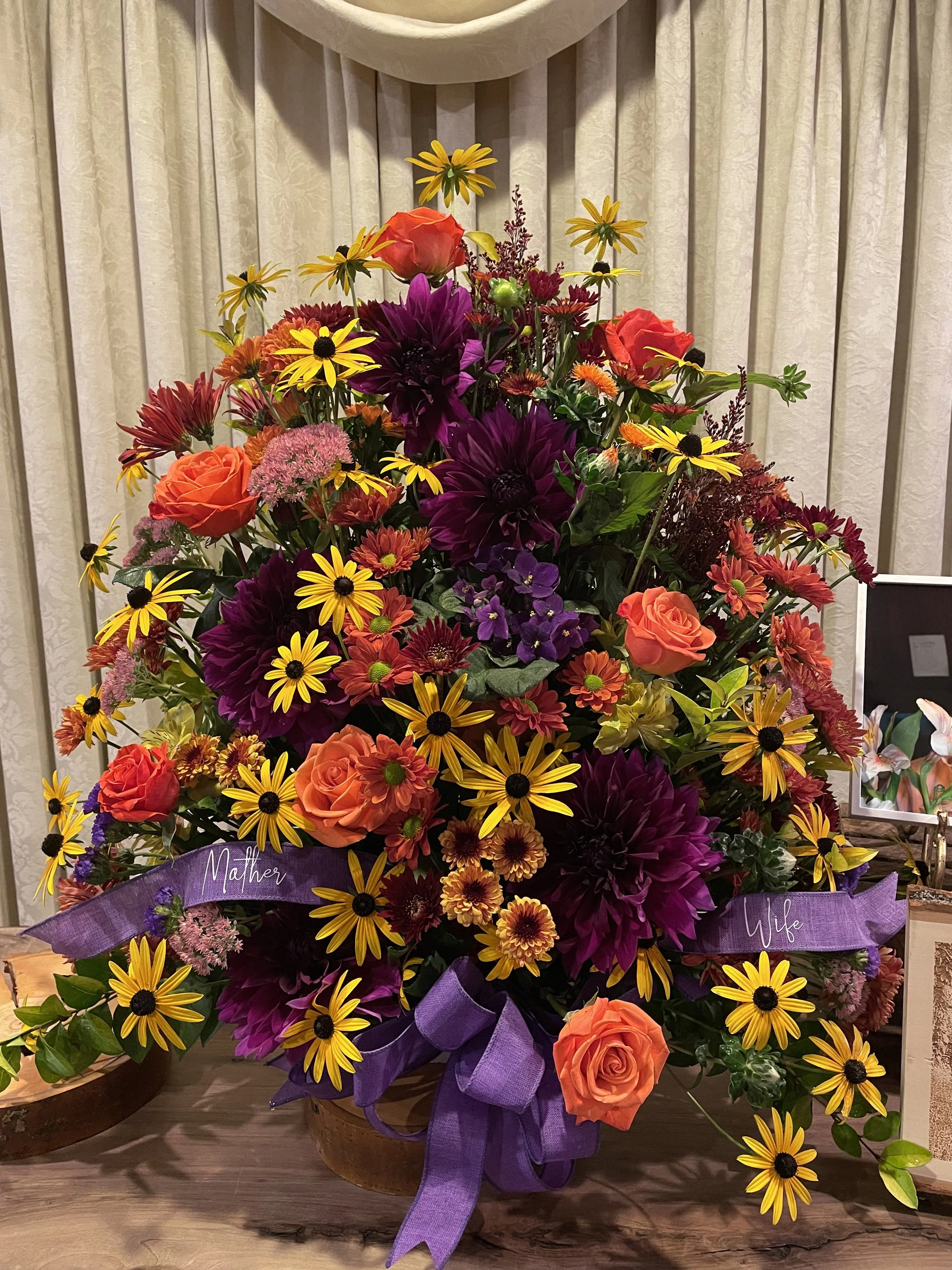 A colorful flower arrangement with orange roses, yellow daisies, purple dahlias, and pink flowers, decorated with purple ribbons labeled 'Mother' and 'Wife'.
