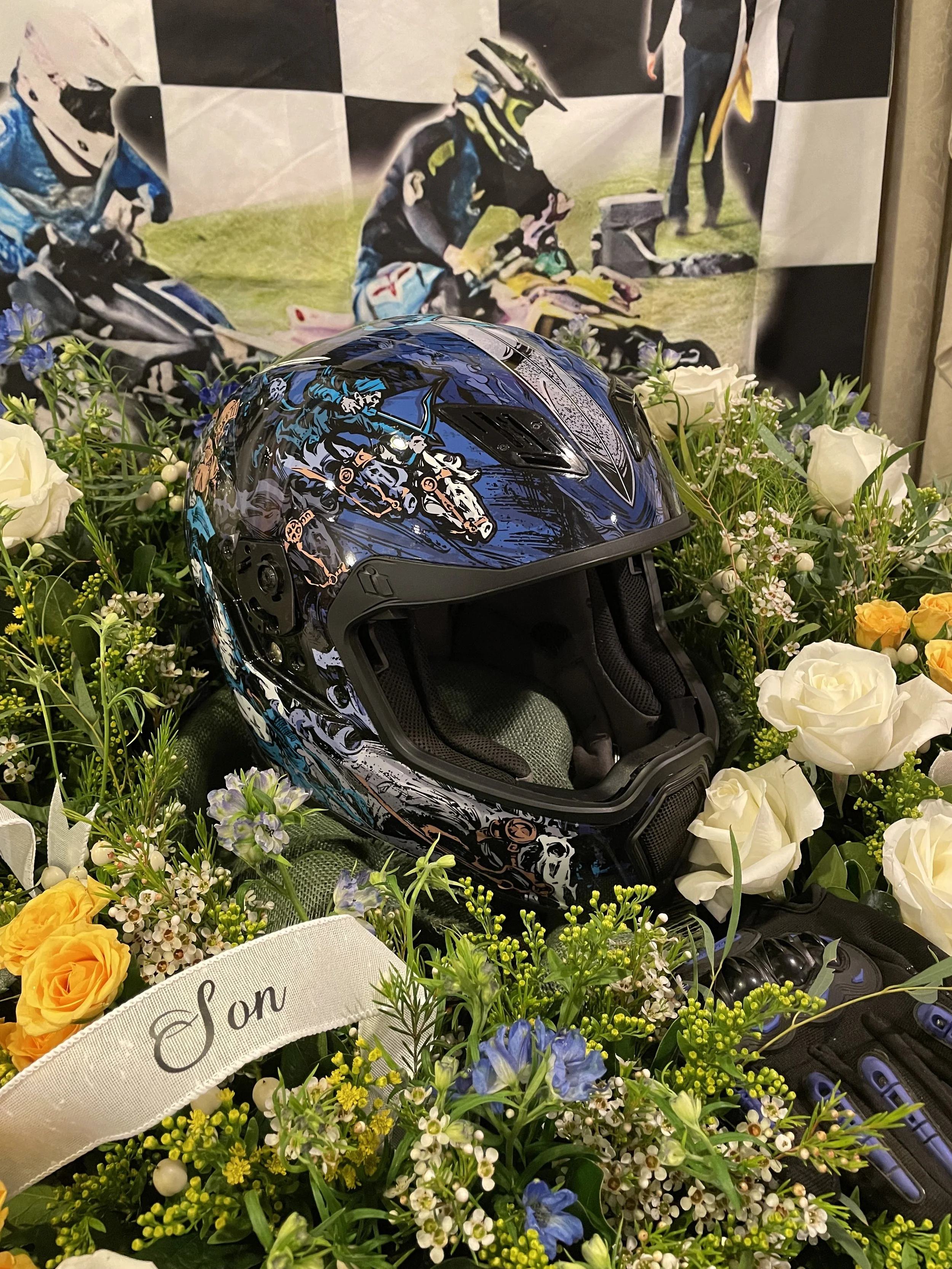 A motorcycle helmet with a colorful graphic design surrounded by a variety of white, yellow, purple, and green flowers and plants. In the background, there is a blurred poster of two motocross racers.