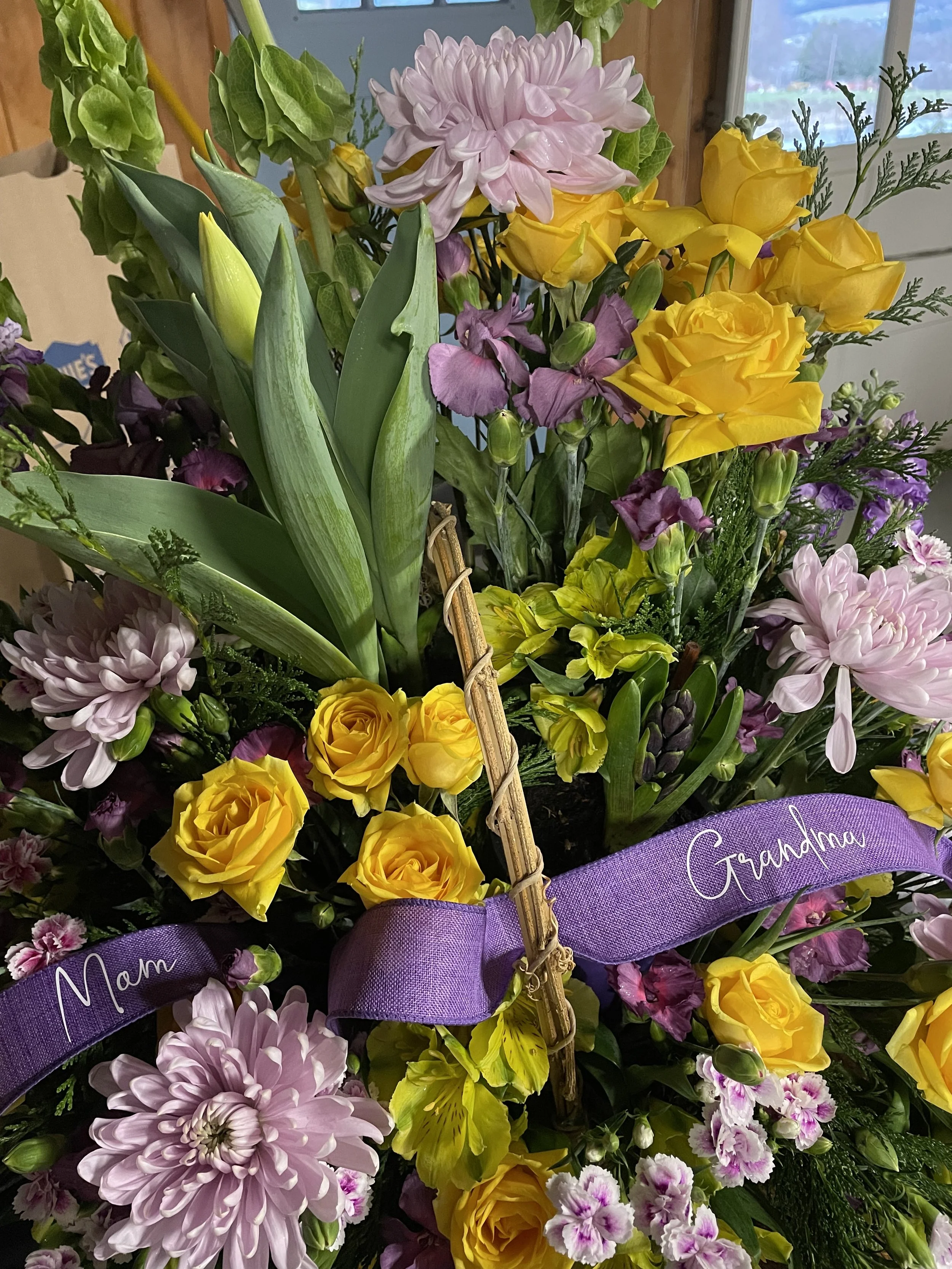 A colorful bouquet of flowers including yellow roses, pink chrysanthemums, purple lisianthus, and green leaves, with a purple ribbon that has "Mom" and "Grandma" written on it.