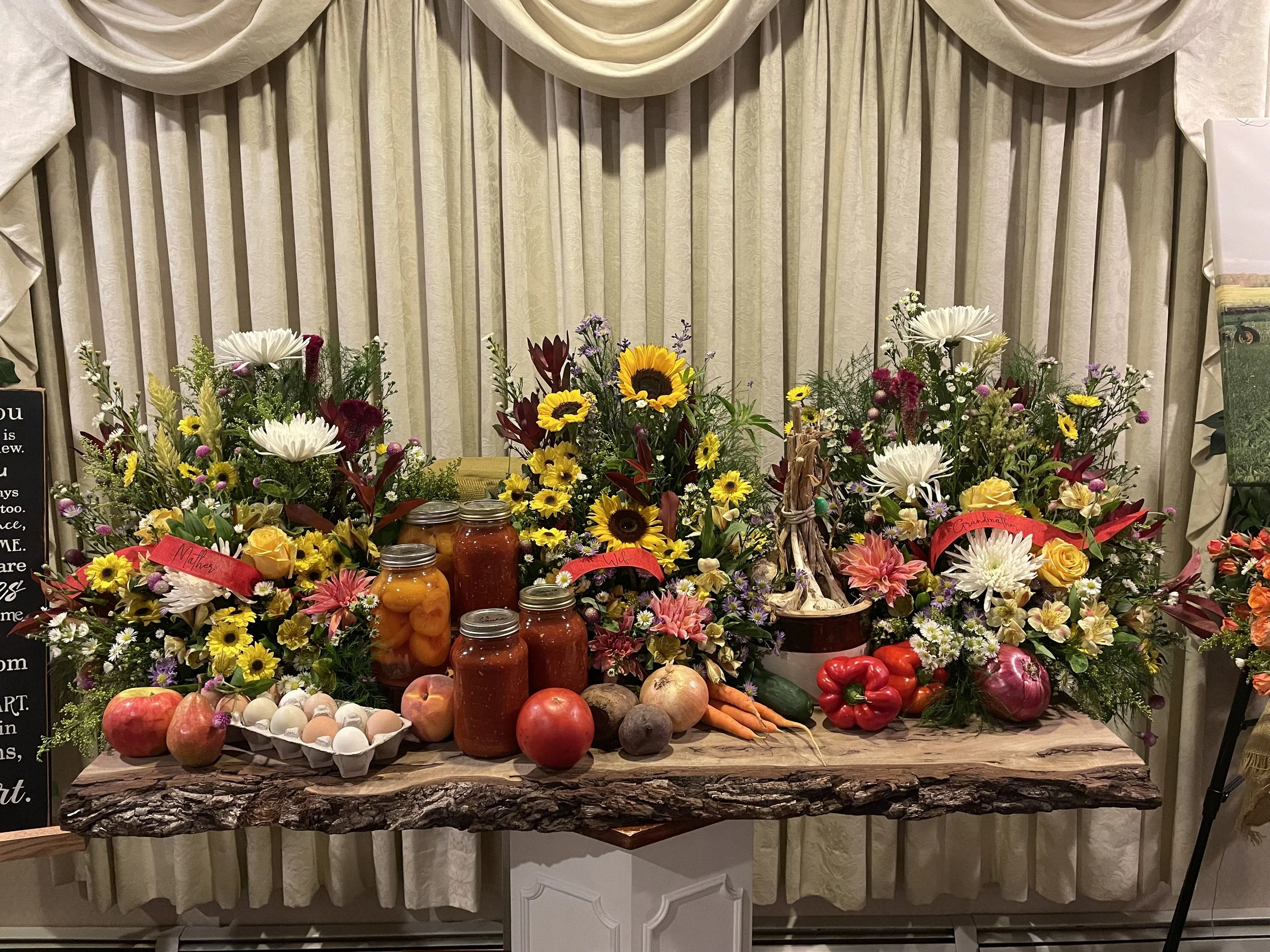 A rustic table covered with a variety of fresh vegetables, fruits, and jars of preserved foods, surrounded by colorful flower arrangements against a cream-colored curtain backdrop.