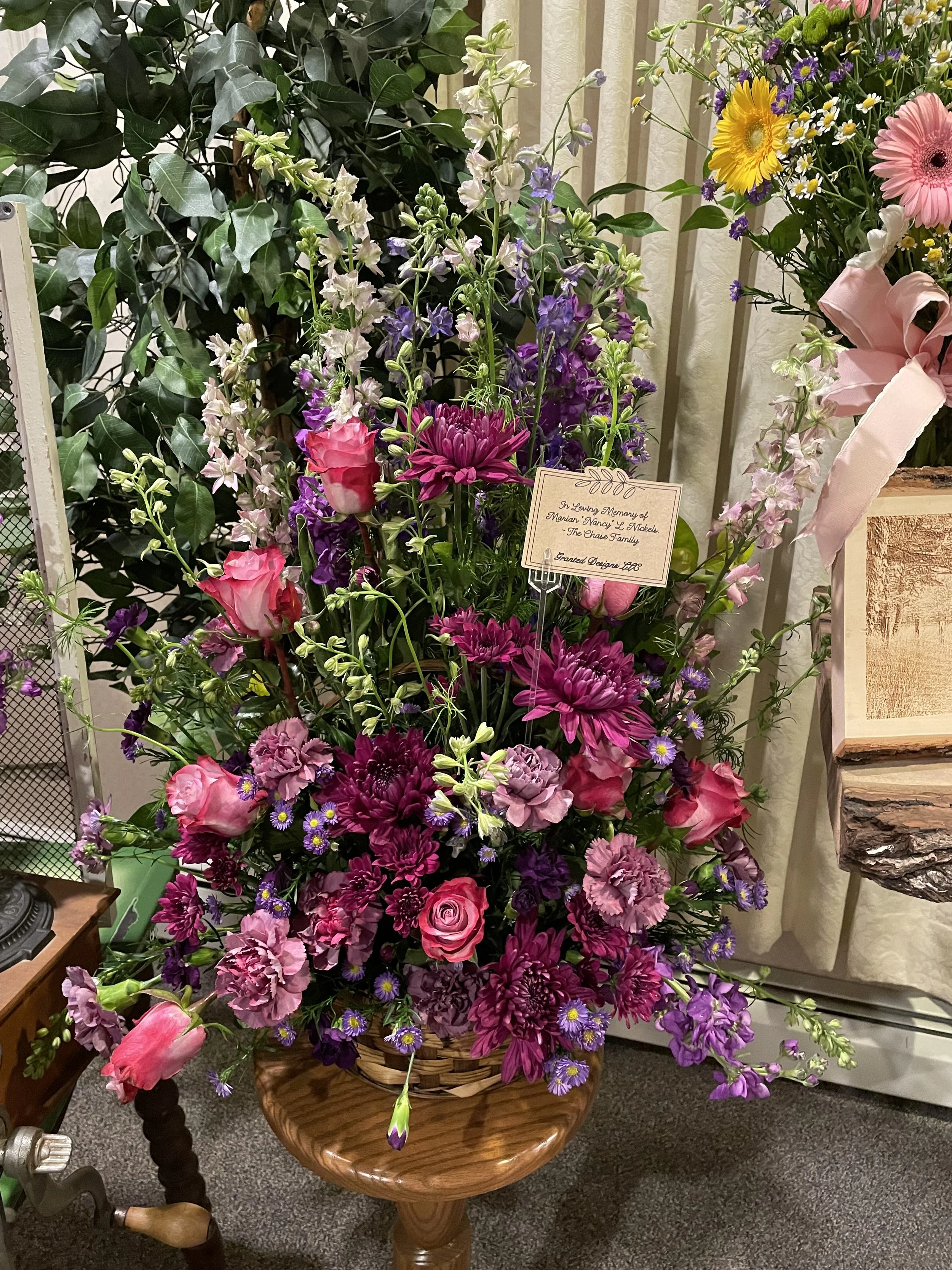 A large floral arrangement in a woven basket on a wooden pedestal, featuring pink roses, purple chrysanthemums, and other purple and pink flowers, with a memorial note attached.