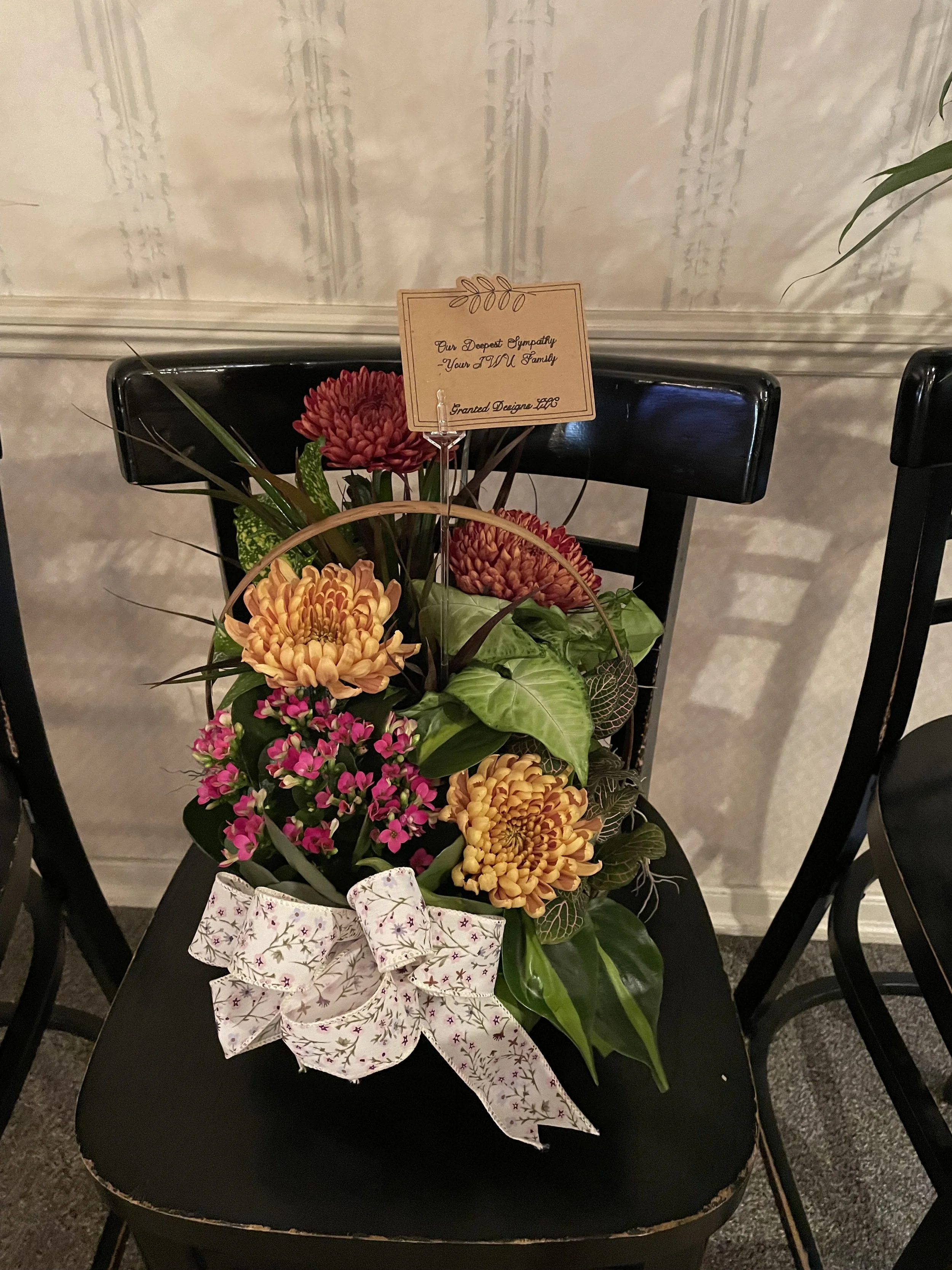 A flower arrangement with large orange and red blooms, small pink flowers, and green leaves, decorated with a white bow with pink stars, placed on a black chair with a note attached.