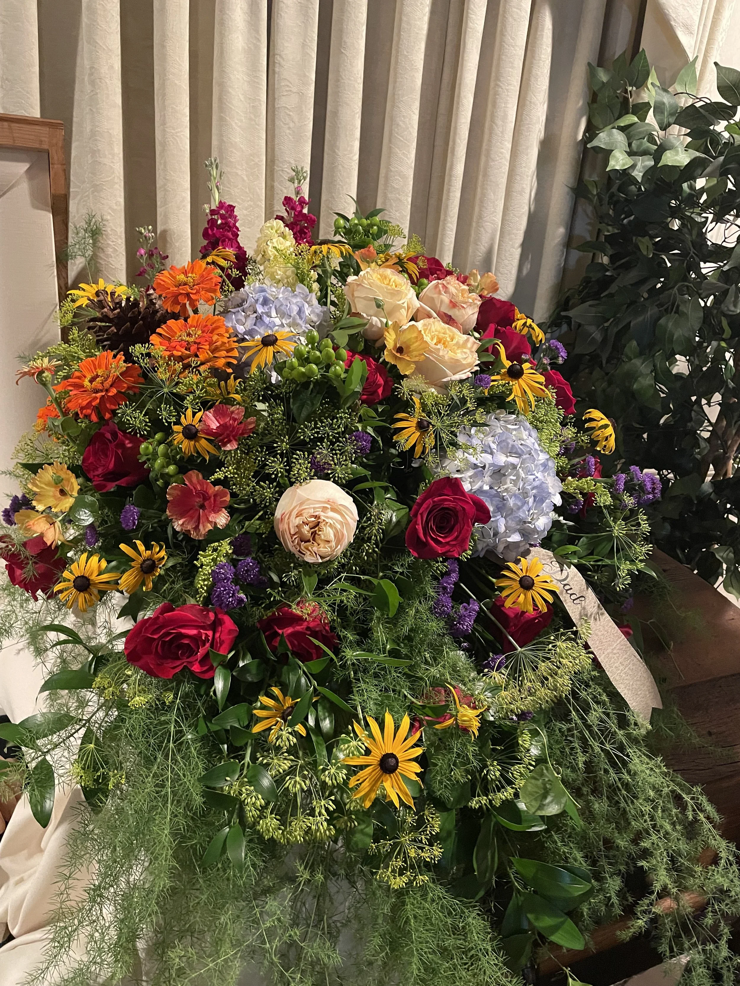 A lush floral arrangement with red roses, peach roses, purple hydrangeas, yellow and orange daisies, and green foliage, placed indoors against cream curtains.