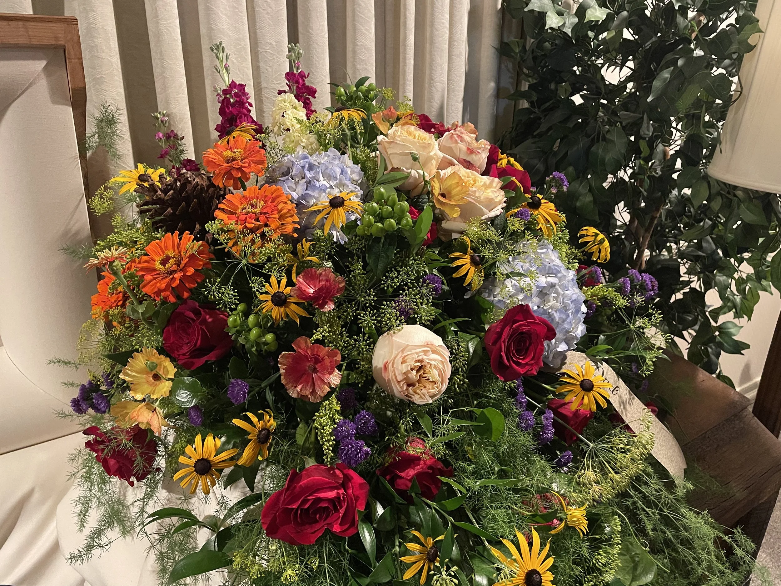 Colorful flower arrangement with roses, hydrangeas, daisies, and other flowers, surrounded by greenery, placed indoors near curtains and a lamp.