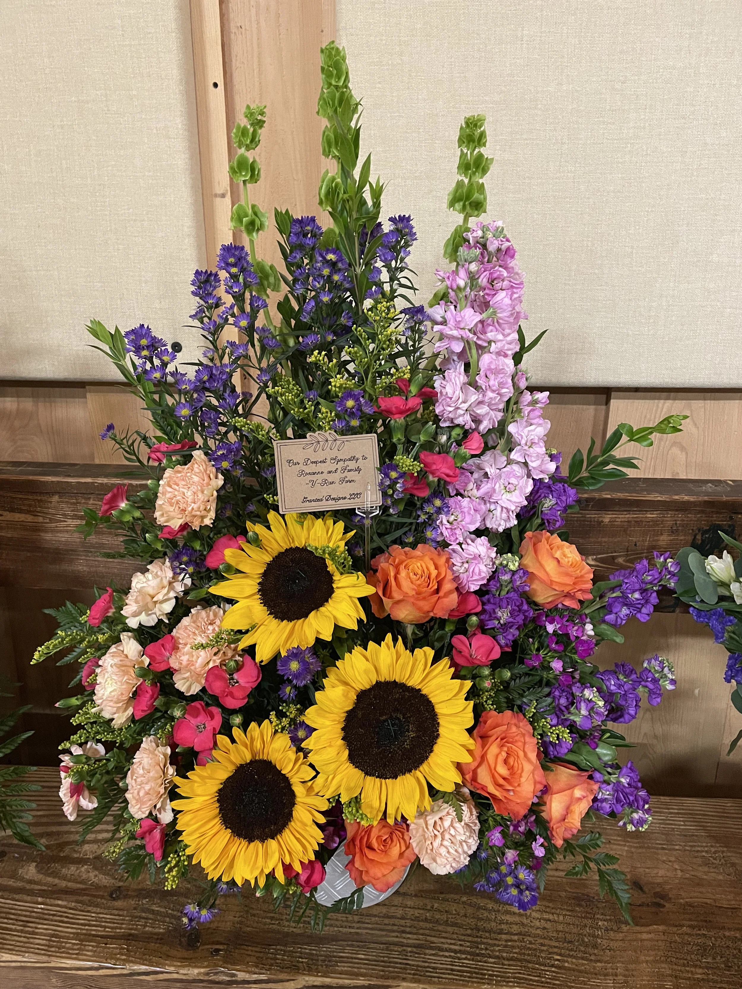 Colorful flower arrangement with sunflowers, roses, carnations, delphiniums, and asters, with a condolence note, on a wooden surface.