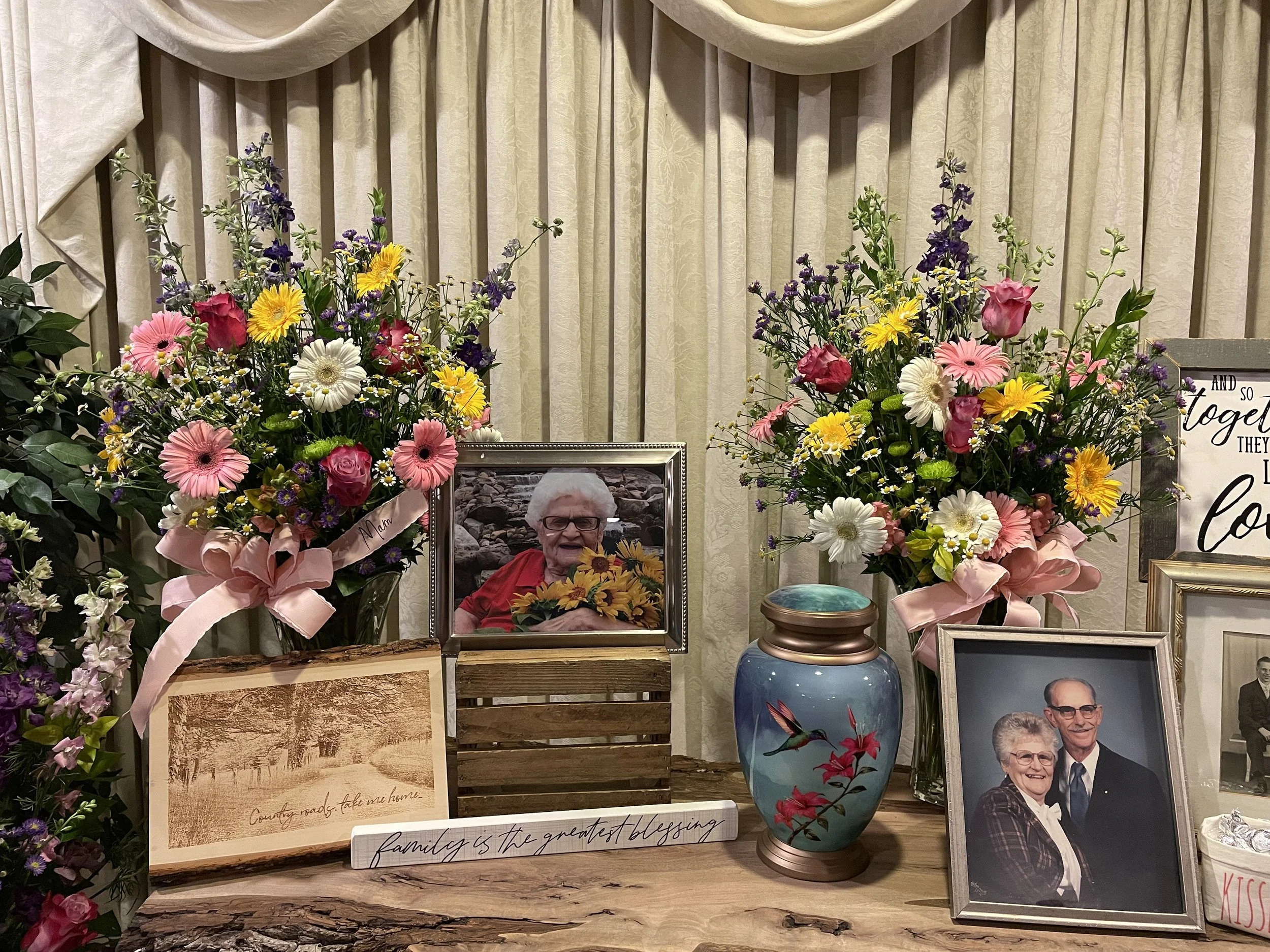 Flower arrangement with pink, yellow, white, purple flowers, framed photographs of elderly people, a decorative vase with birds and flowers, and signs with gratitude messages on a wooden table.