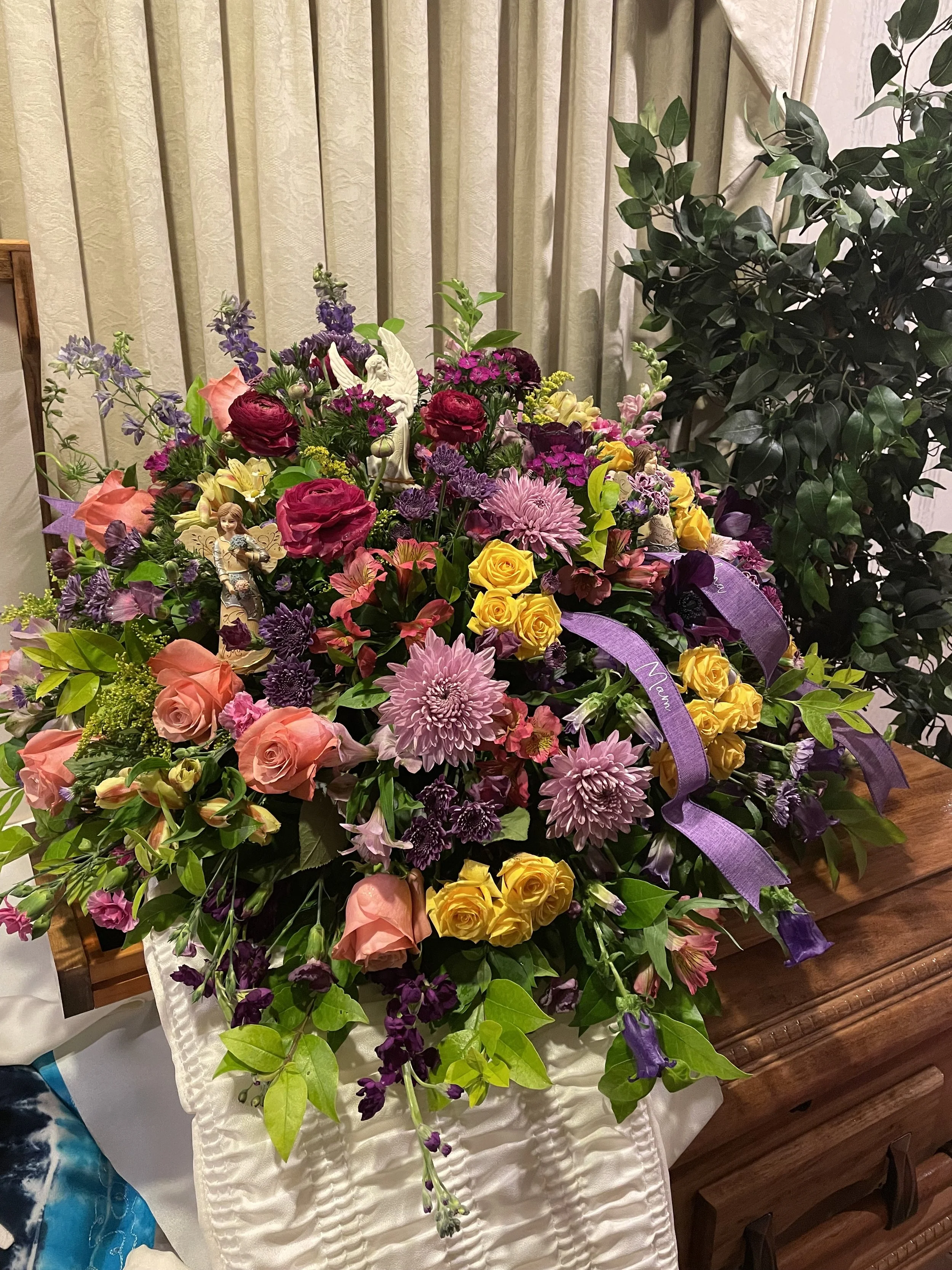 Colorful flower arrangement with pink, purple, yellow, and peach flowers, including roses, chrysanthemums, and other blooms, accented with ribbons and greenery, placed on a wooden table.