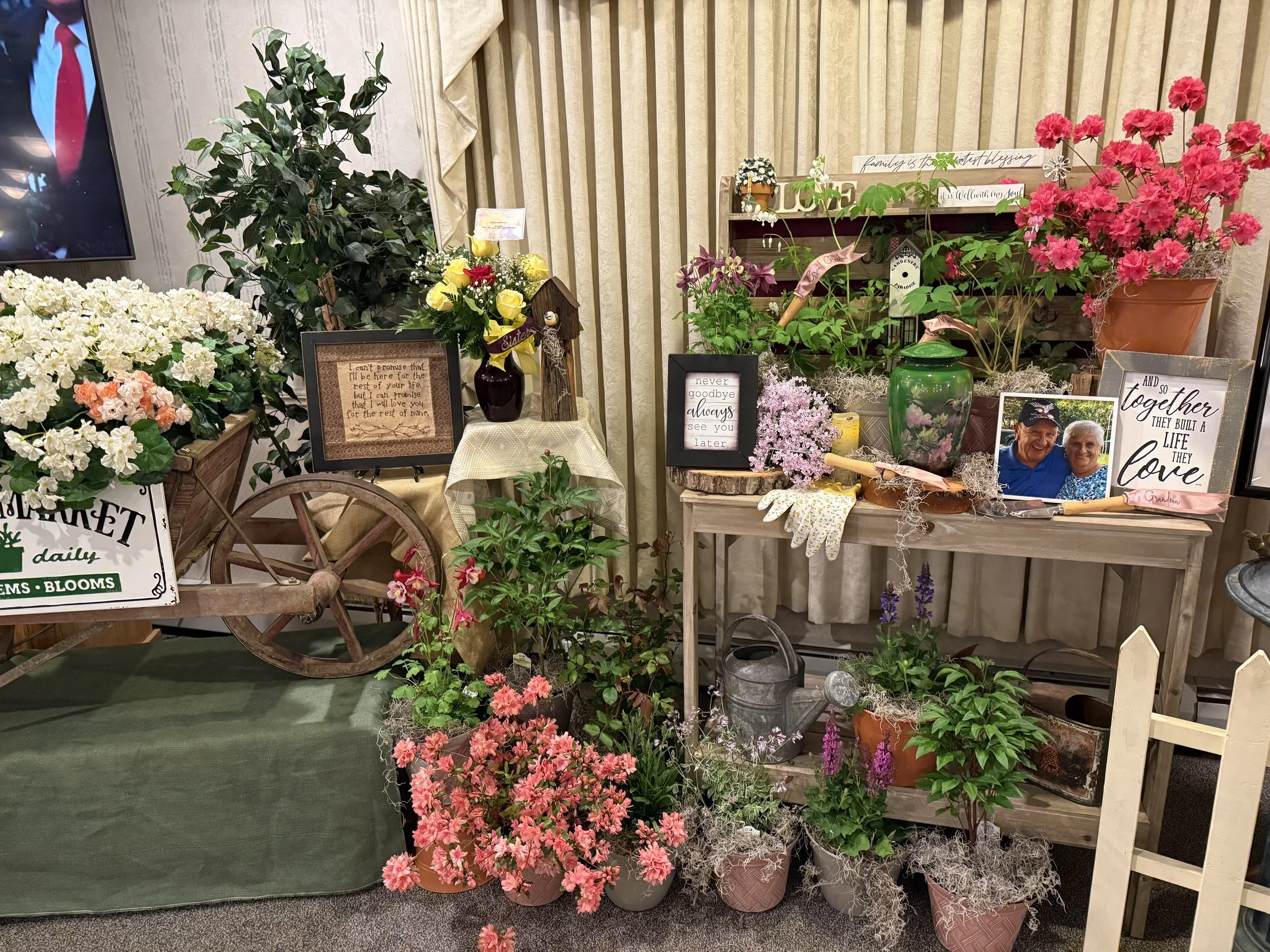 Display of various potted and cut flowers, framed photos, and decorative signs on tables and a cart, with a green curtain and beige curtains in the background.