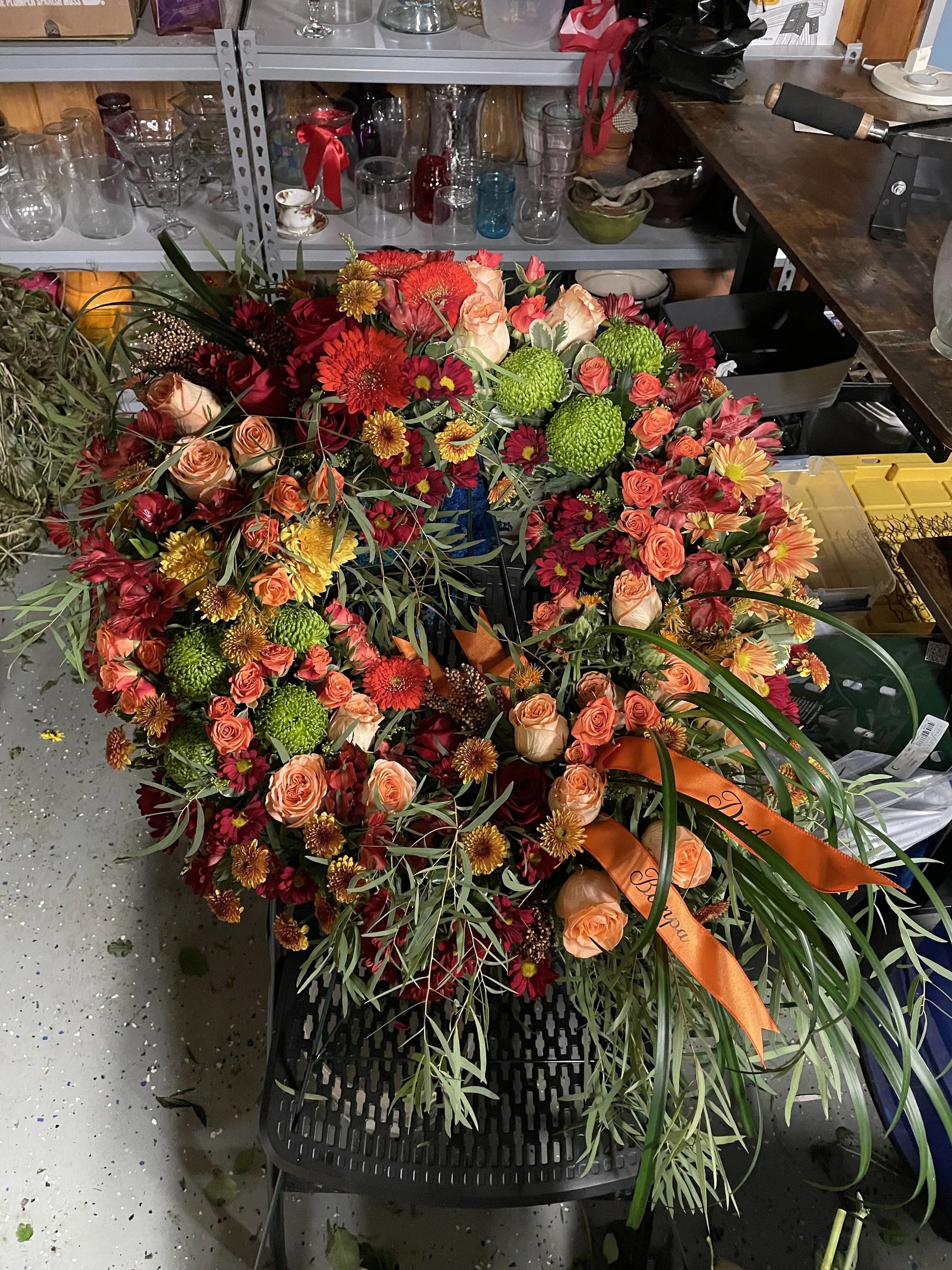 A large, colorful flower arrangement with peach roses, red and orange daisies, and green pom-pom flowers, accented with greenery, placed on a black chair in a storage room or workshop.