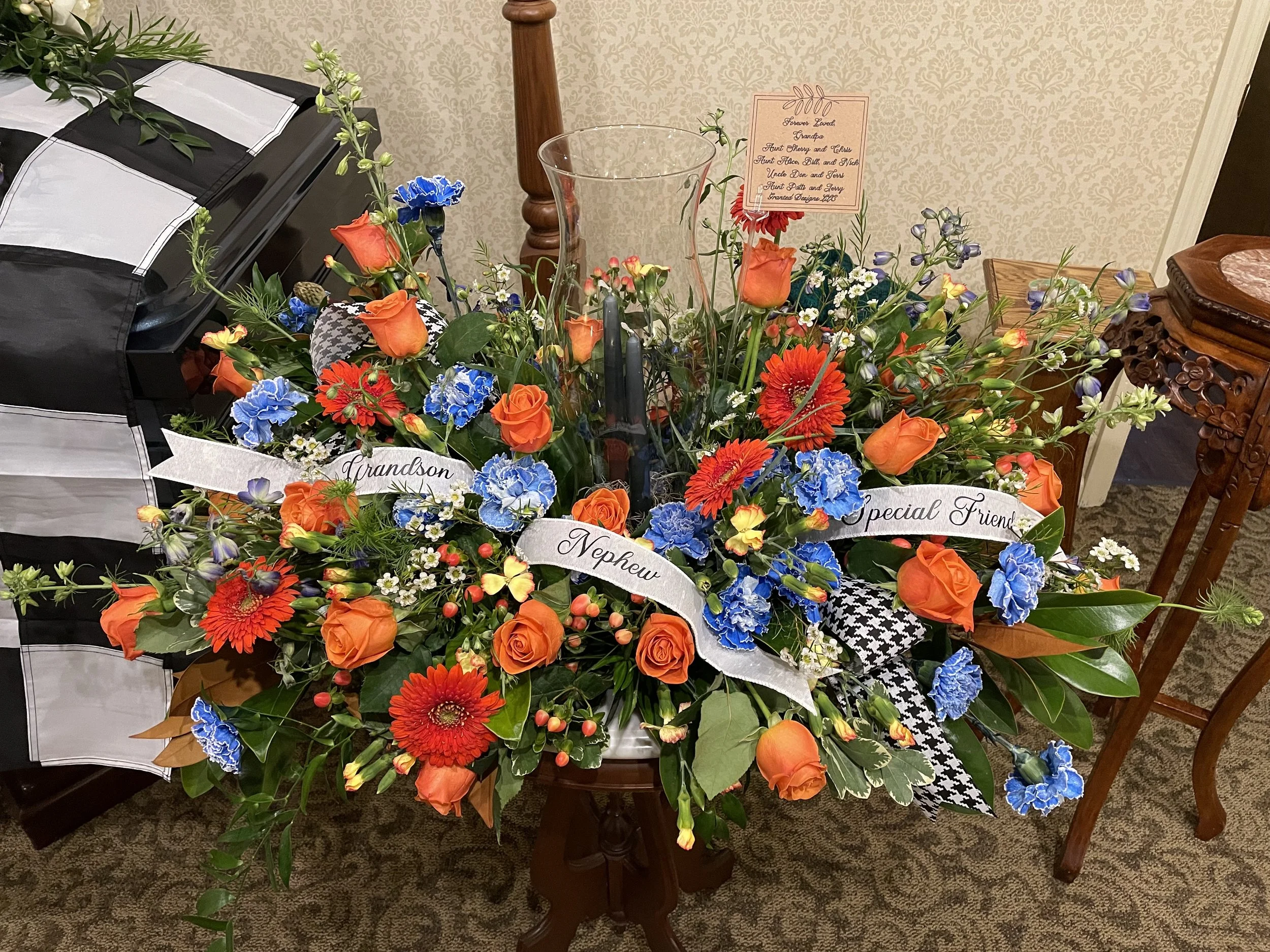 A large floral arrangement with orange roses, red gerbera daisies, blue carnations, and greenery, set on a small wooden table in a room. It includes memorial ribbons with the words "Grandson," "Nephew," and "Special Friend." There is a glass vase, a