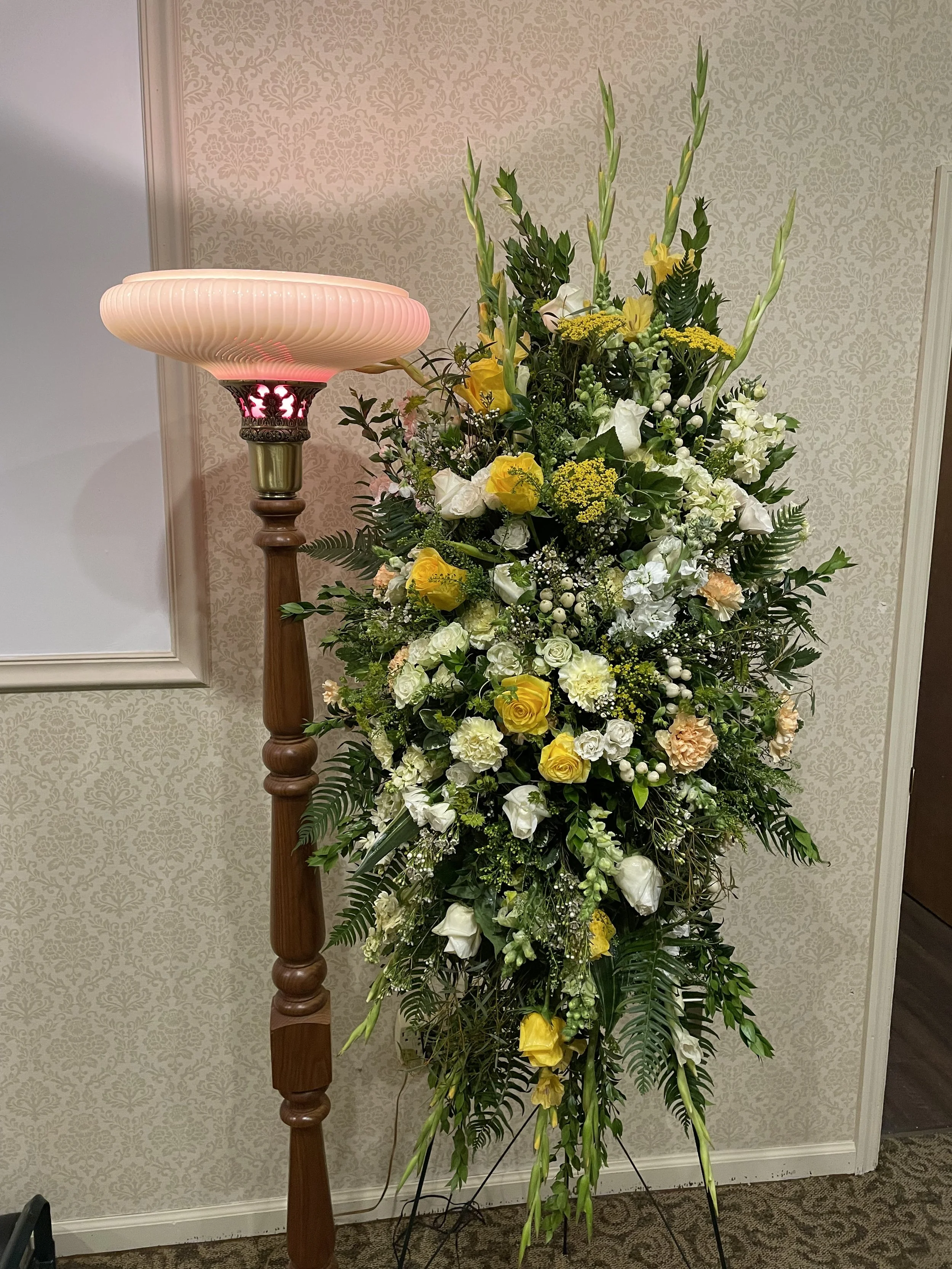 Large floral arrangement with yellow and white flowers, greenery, and tall gladiolus, next to a tall wooden floor lamp with a pink glass shade.