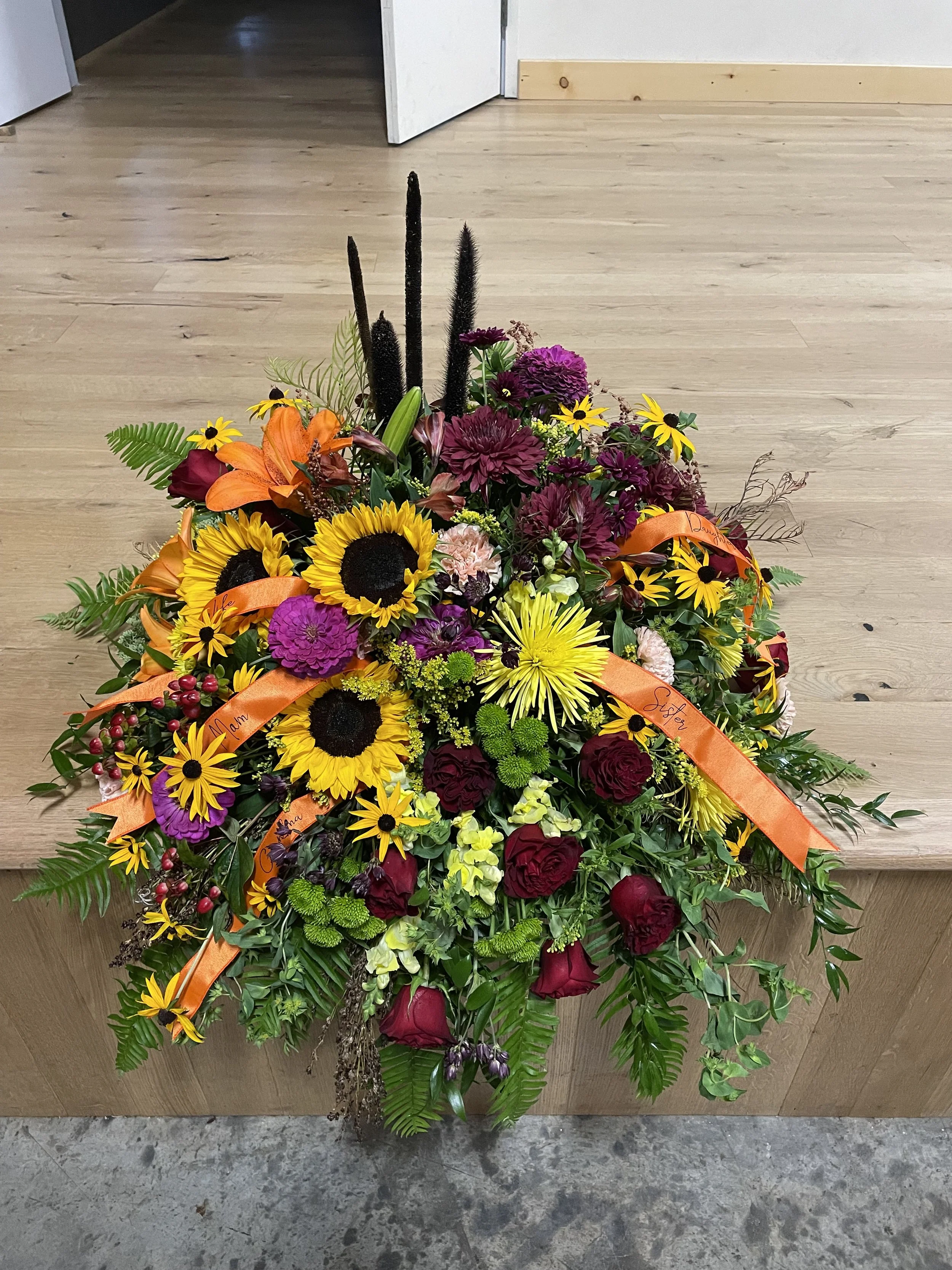 A colorful floral arrangement with sunflowers, purple carnations, orange lilies, deep red roses, yellow daisies, and green foliage placed on a wooden surface.