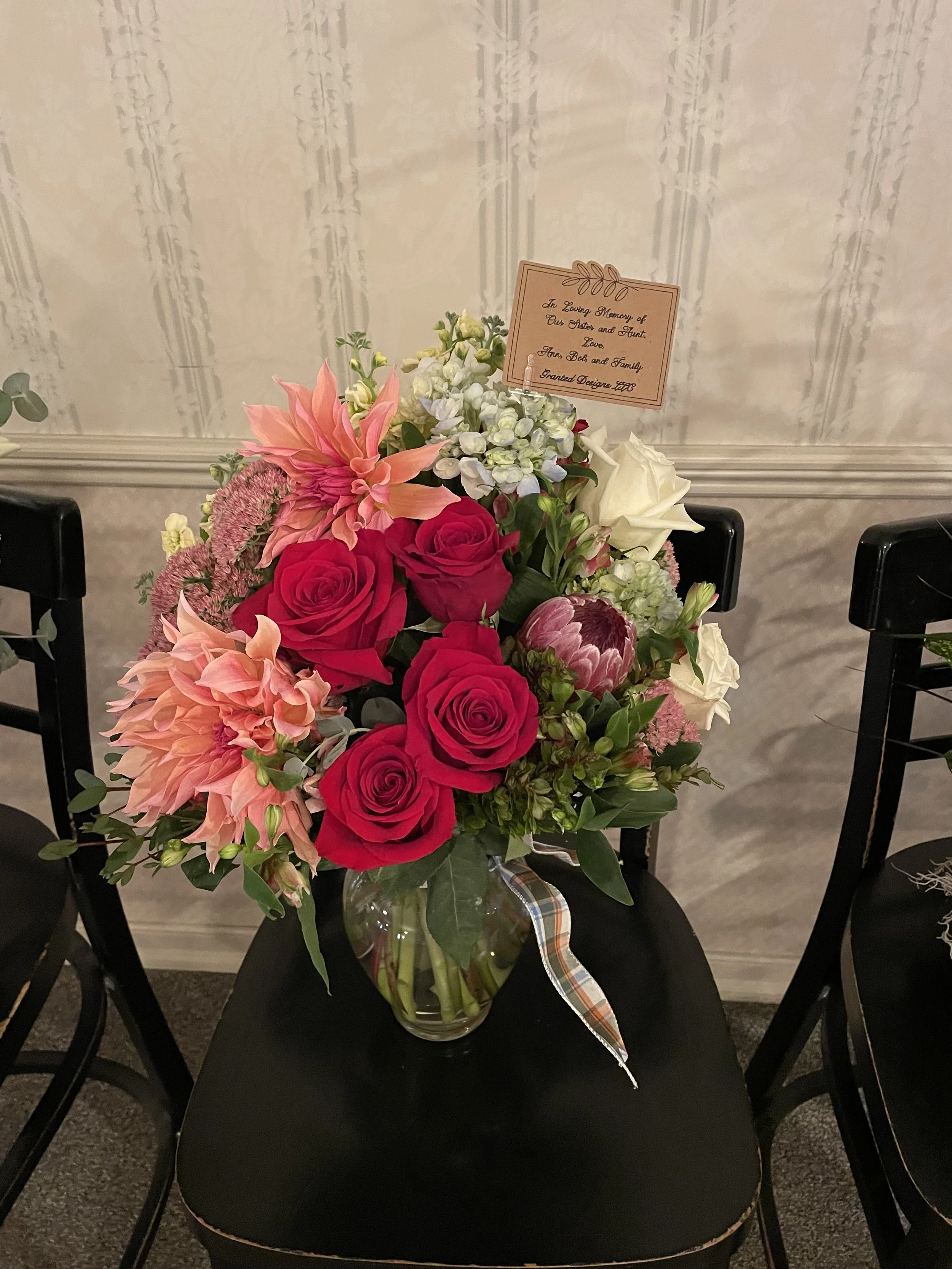 A bouquet of pink roses, peach dahlias, white roses, and other assorted flowers in a clear glass vase on a black chair. A note attached to the bouquet reads, 'In Loving Memory of The Elder and Heart. Love, Tom Bob and Family, Grand Design 2022.'